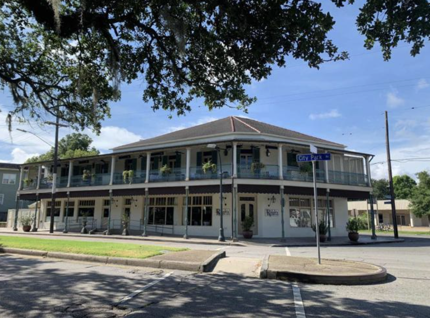 Ralph Brennan purchases the historic two-story building and reopens Ralph's on the Park in 2003, located at an intersection on a sunny day. There is a street sign that reads 'City Park' and a bright blue sky with some clouds.