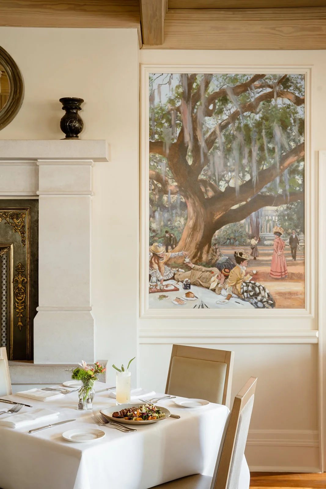 A dining room with a white tablecloth featuring plates, utensils, a small floral centerpiece set for one. Behind it, a large painting depicts a park scene with people lounging under a large tree, some sitting on a picnic blanket and others walking.