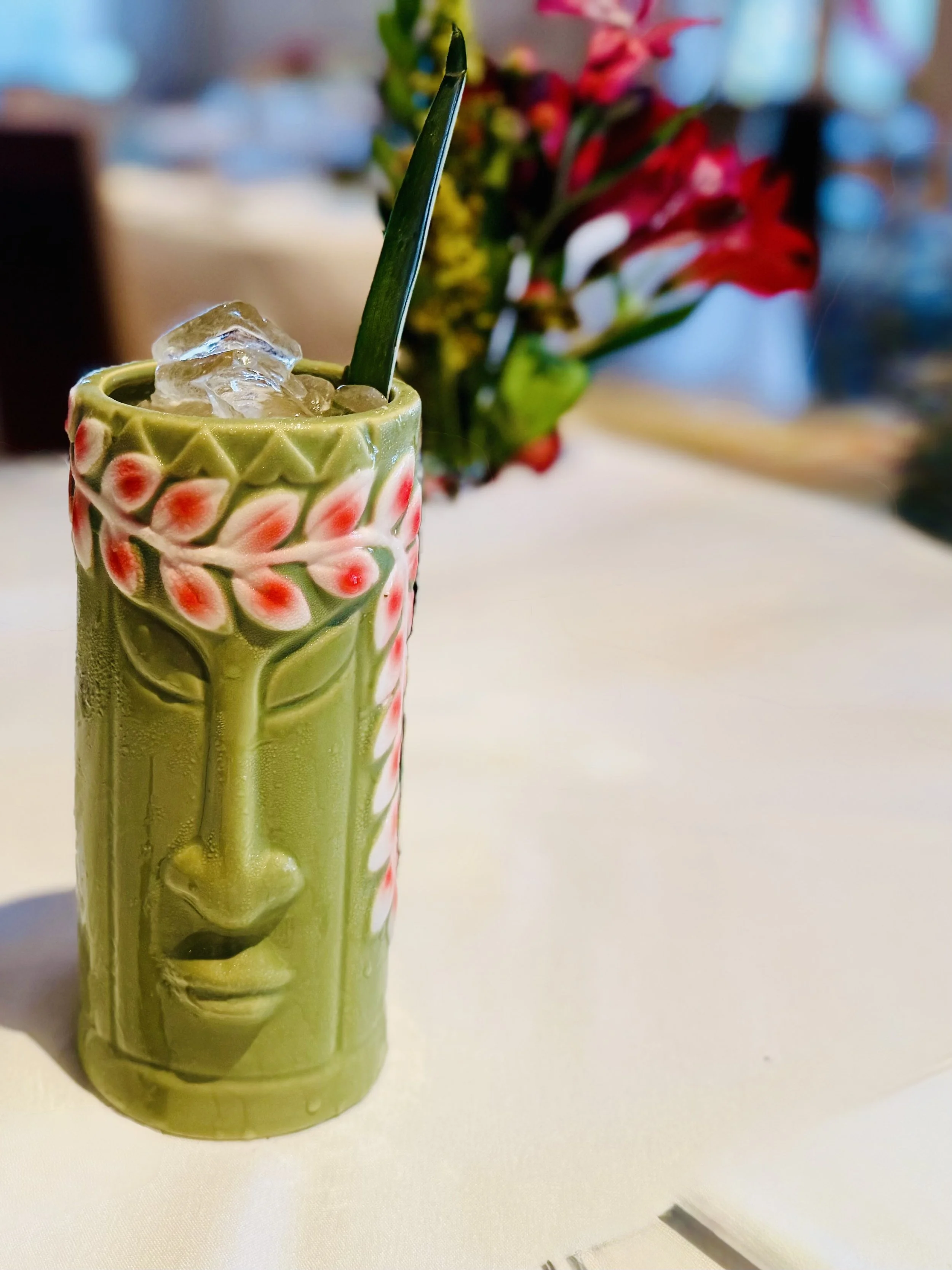 A tiki-style ceramic cup with a tropical face design, filled with ice and a drink, garnished with a green straw, in front of a blurred background of colorful flowers.