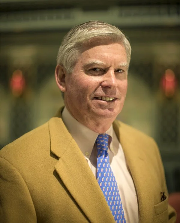 Portrait of a middle-aged man with gray hair, wearing a tan blazer, white dress shirt, and a blue patterned tie, standing indoors with blurred background.