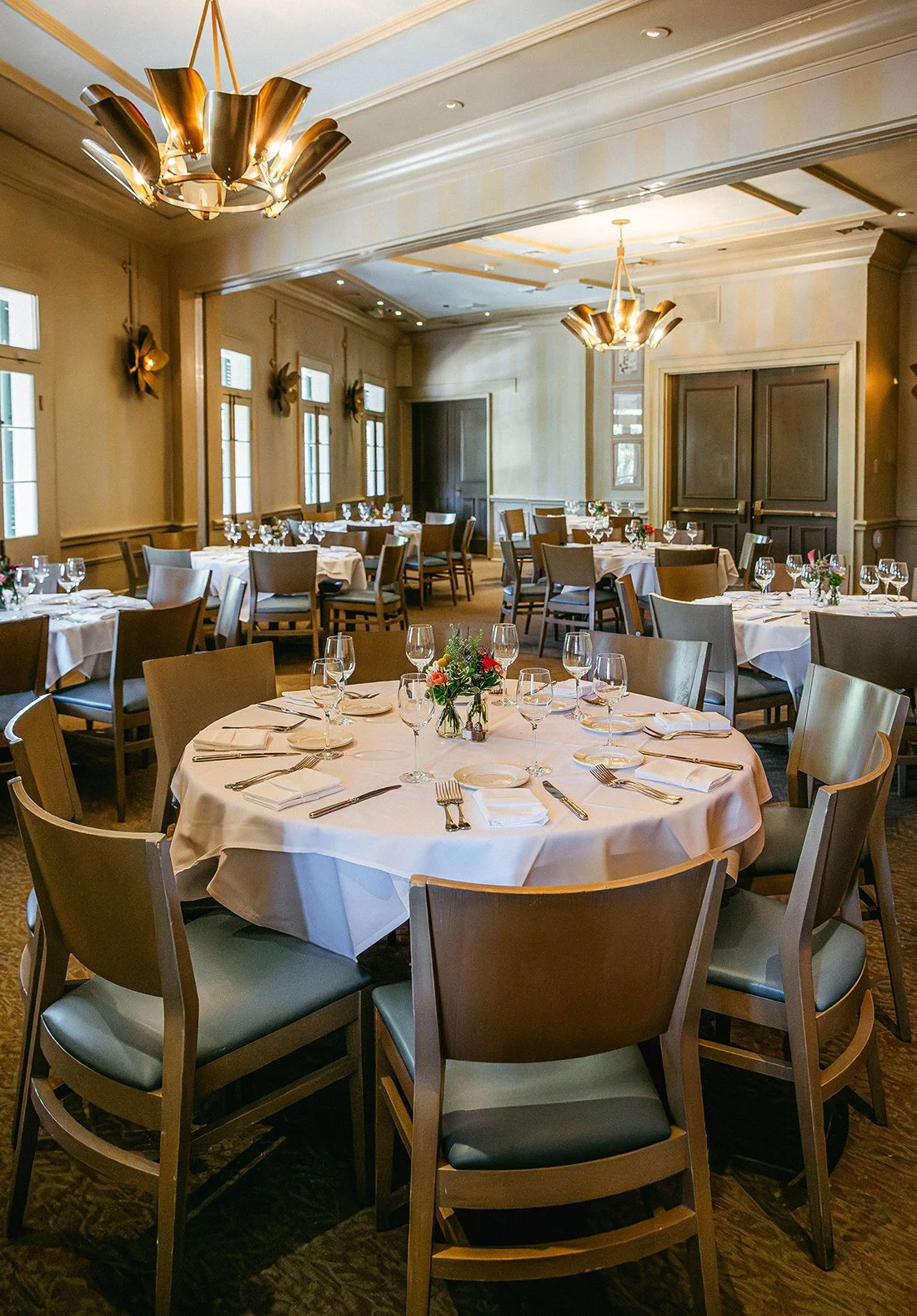 Elegant banquet room with round tables covered in white tablecloths, set with glasses, plates, silverware, white napkins, and small floral centerpieces, illuminated by warm lighting.