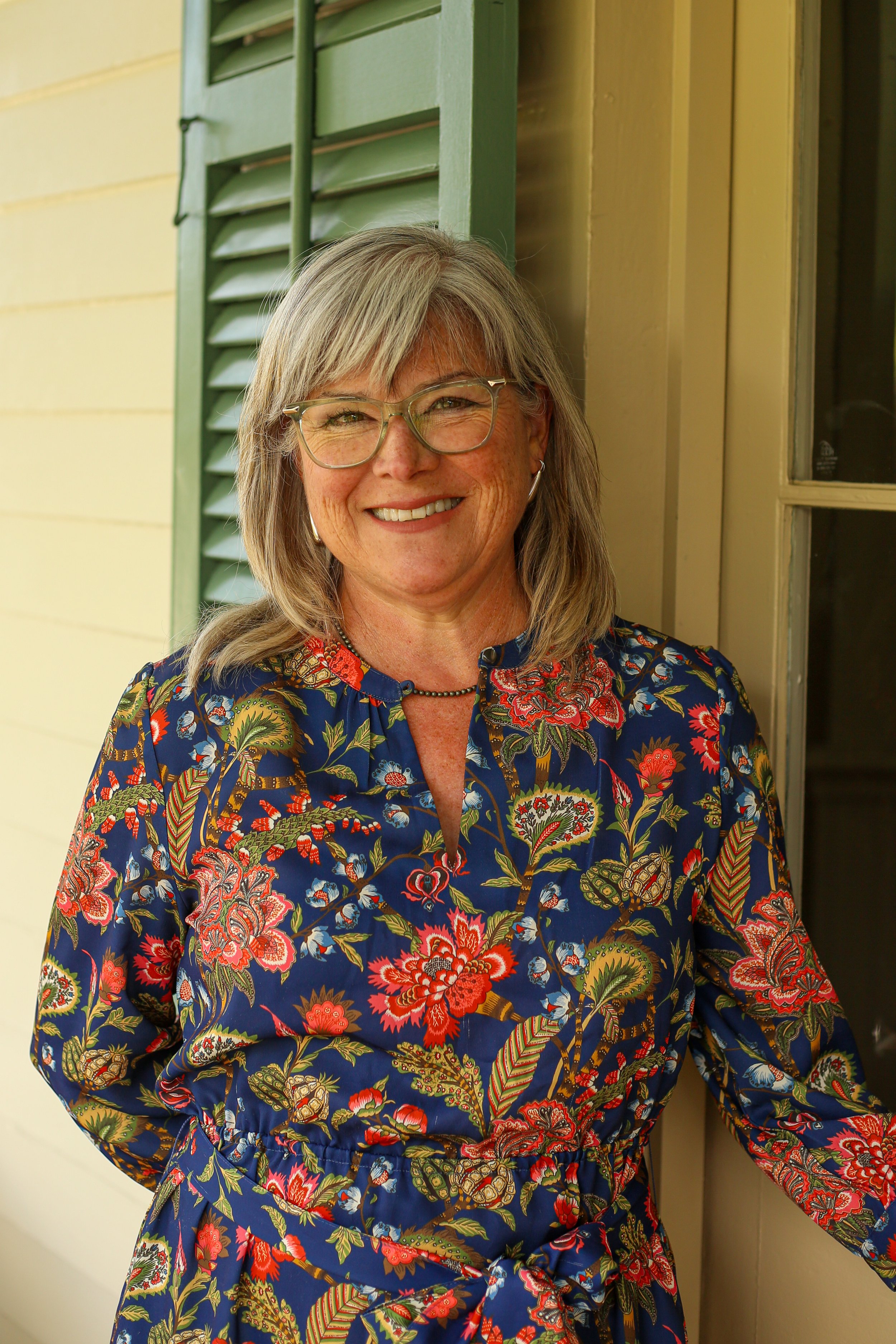 A smiling middle-aged woman with gray hair, wearing a navy blue sleeveless blouse with a bow, standing in a doorframe with green shutters.
