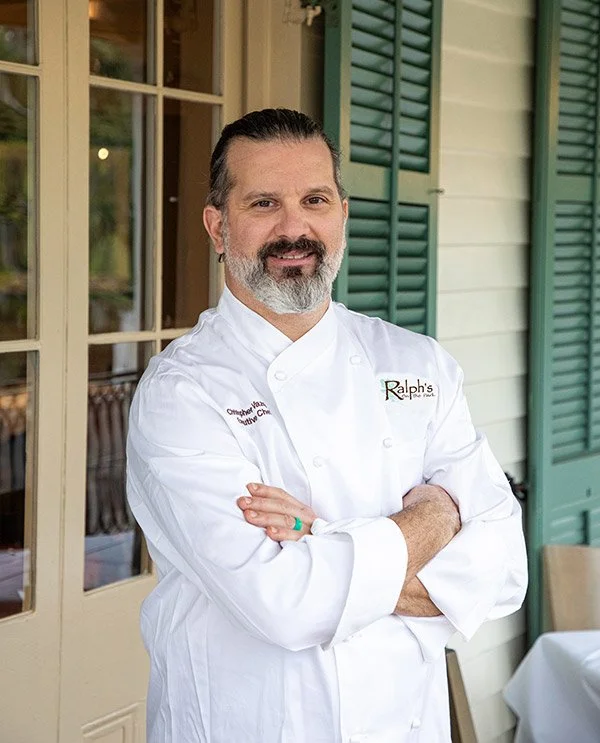 A man in a white chef's coat with folded arms, standing outside a building with green shutters and a window.