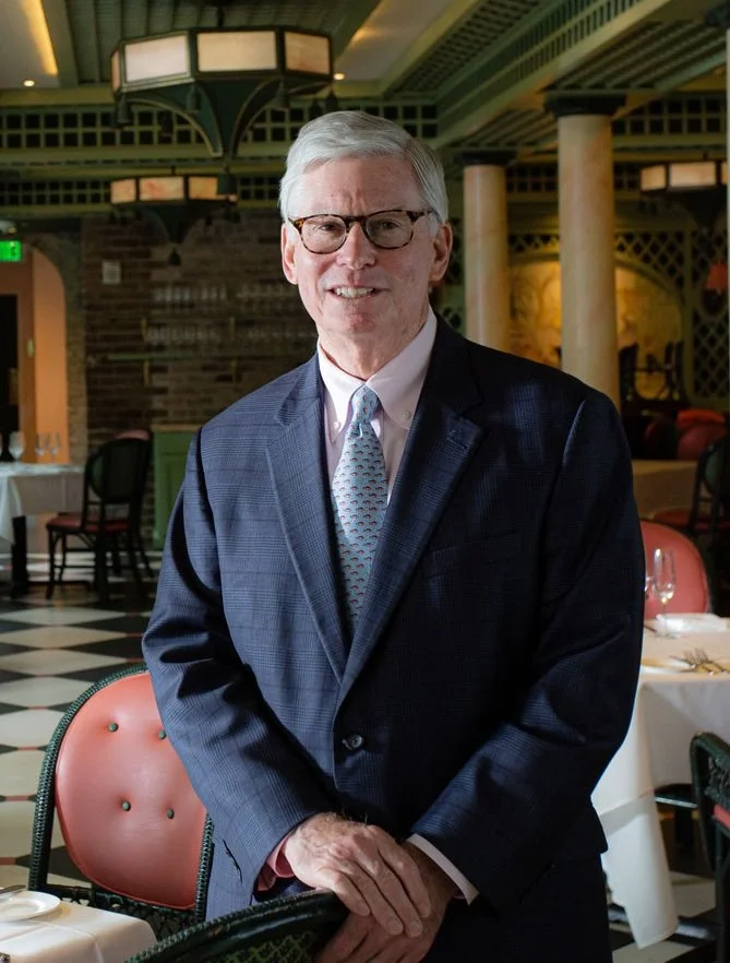 Portrait of a middle-aged man with gray hair, wearing a tan blazer, white dress shirt, and a blue patterned tie, standing indoors with blurred background.