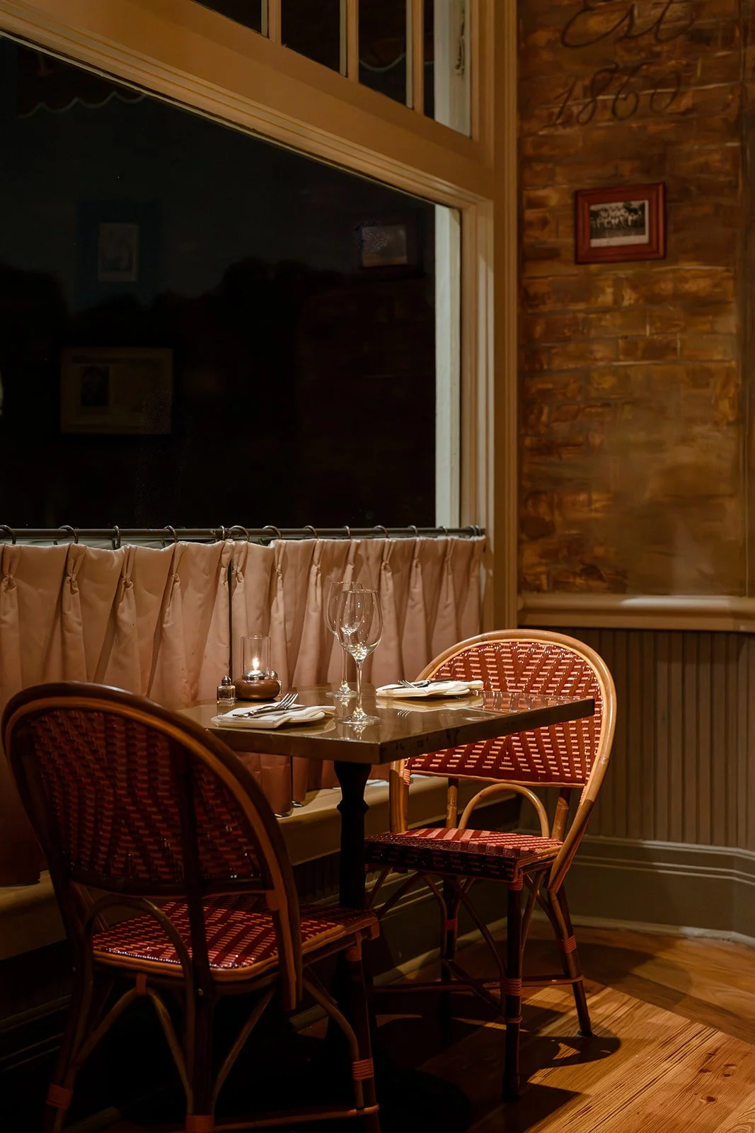 A dimly lit restaurant table set with a candle, wine glass, napkins, and cutlery, next to a window with beige curtains and wooden walls.