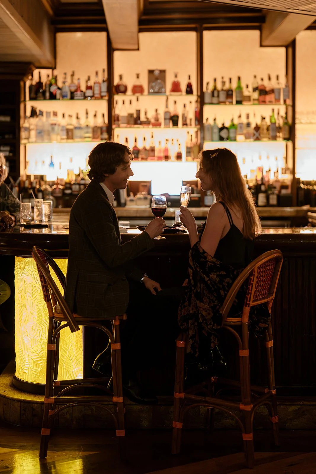 A man and woman enjoying drinks at a bar with warm lighting, shelves of liquor bottles in the background, and two empty chairs in front of them.