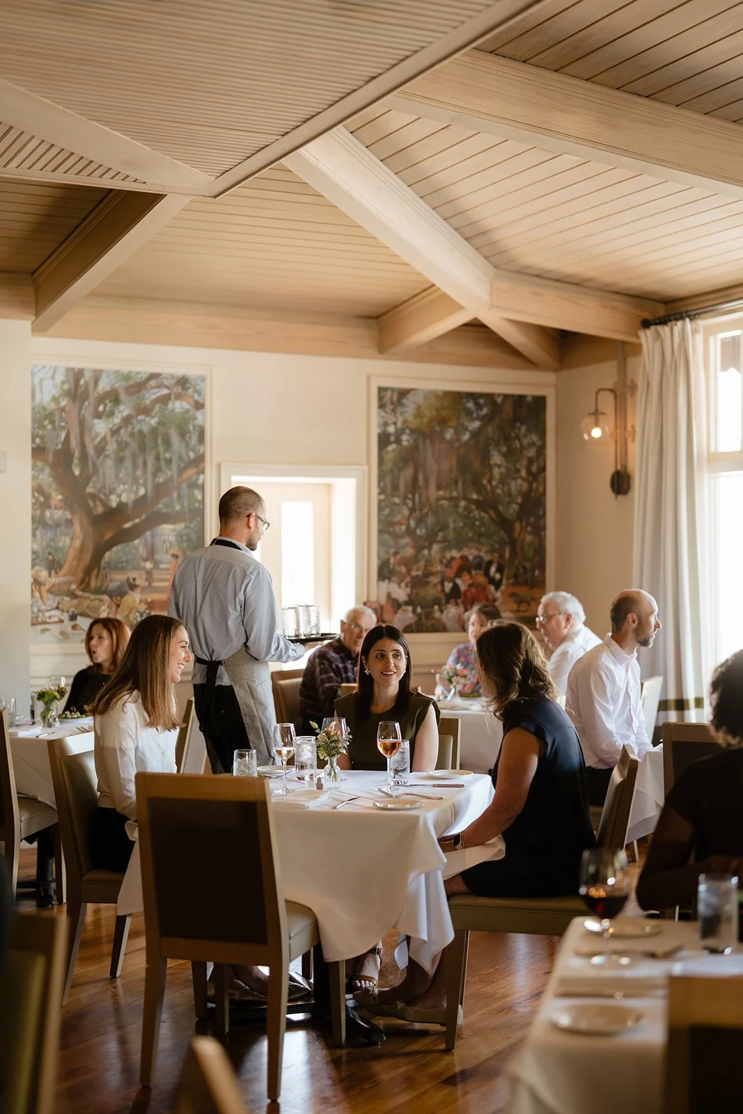 People dining in a restaurant with artwork on the walls, natural light, and a waiter serving drinks.