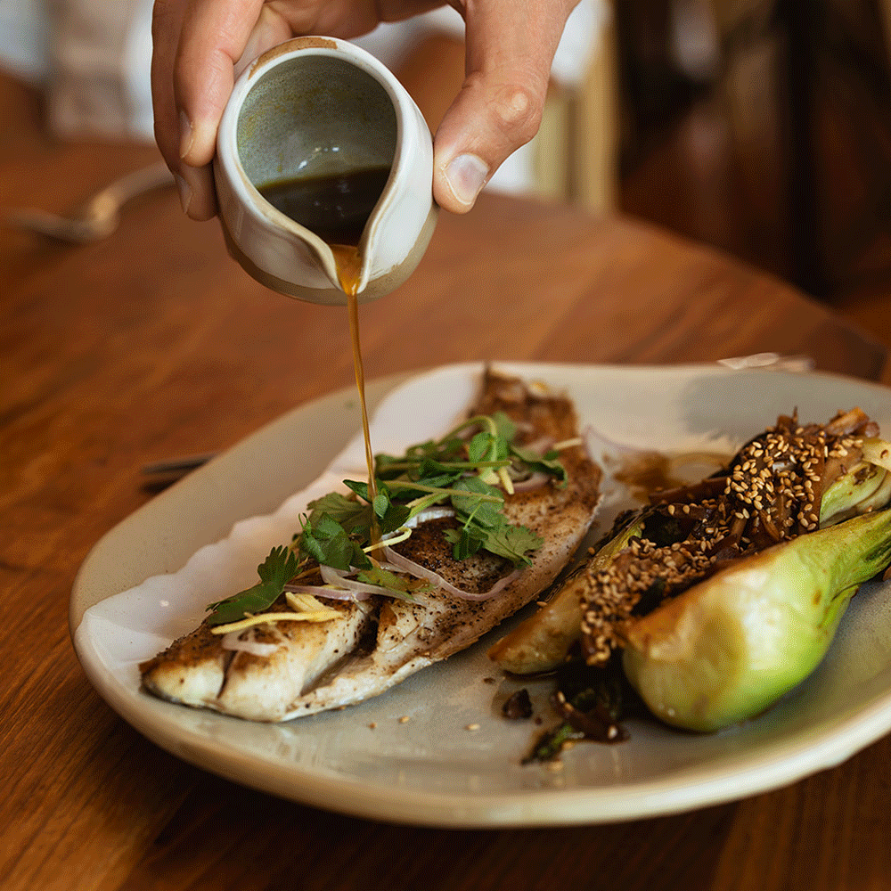 Person pouring sauce onto grilled fish with herbs, accompanied by roasted stuffed vegetable on a ceramic plate