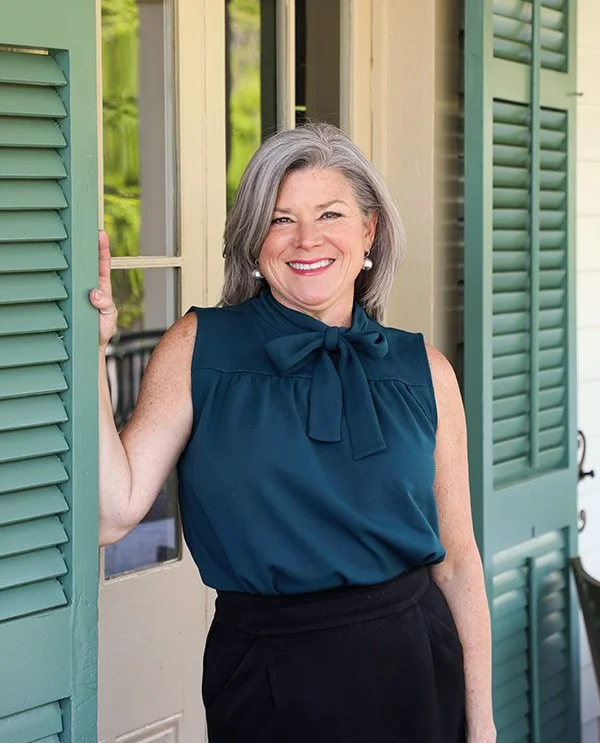 A smiling middle-aged woman with gray hair, wearing a navy blue sleeveless blouse with a bow, standing in a doorframe with green shutters.