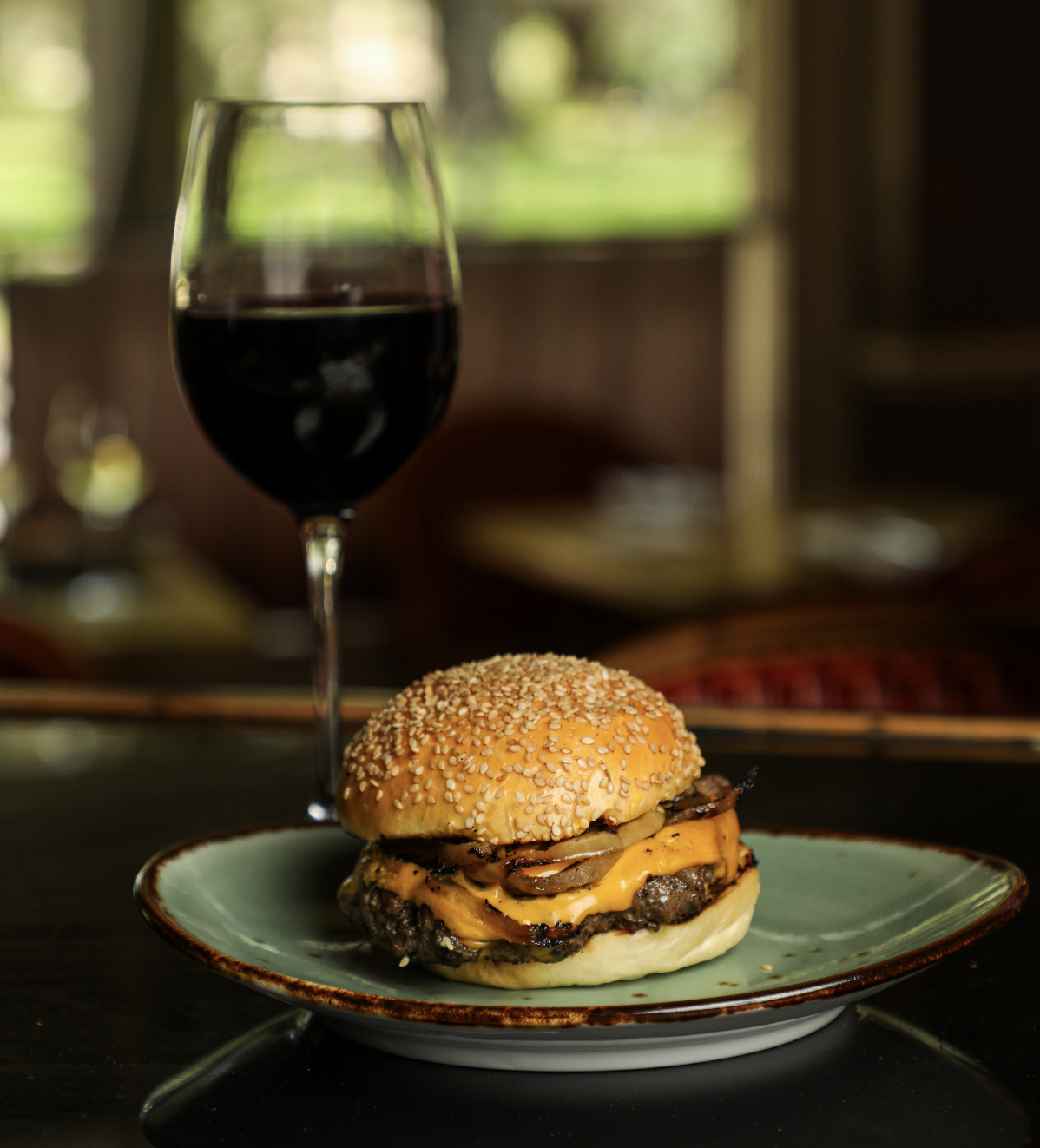 A cheeseburger with a sesame seed bun, melted cheddar cheese, and beef patty on a green plate. A glass of red wine in the background.