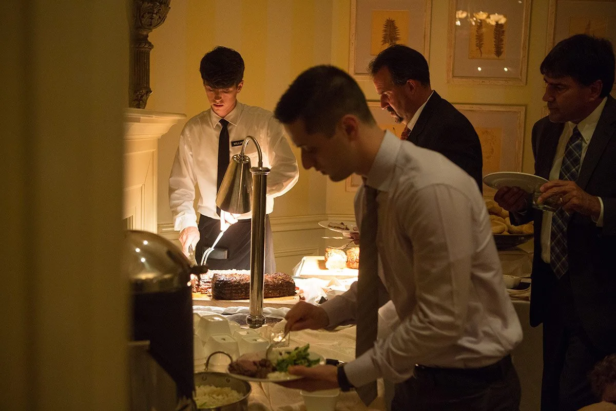People serving themselves food at a buffet in a dimly lit room, with some plates and various dishes on the table.