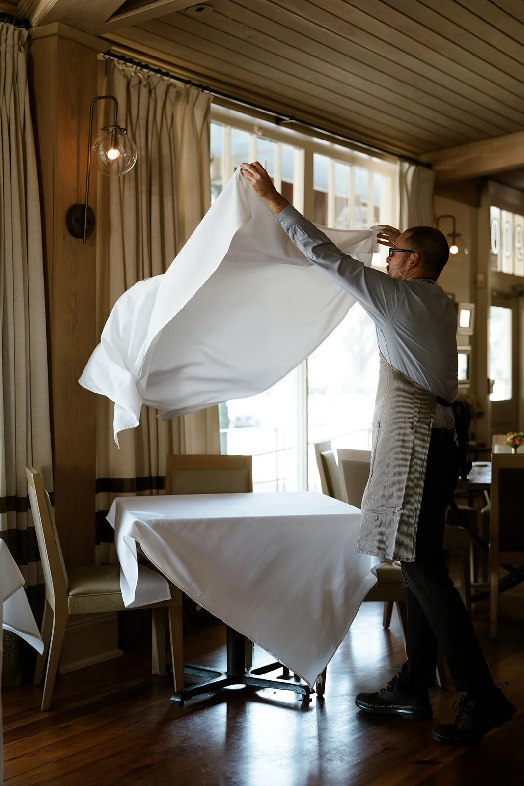 A man wearing glasses and an apron is putting on a white cloth, possibly a tablecloth, in a well-lit room with wooden walls and curtains. There is a table with a white tablecloth in front of him and chairs around it.