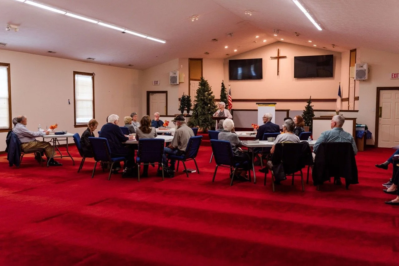 A group of elderly people dining at a long table decorated with Italian flags and tablecloths in red, white, and green, in a community room.