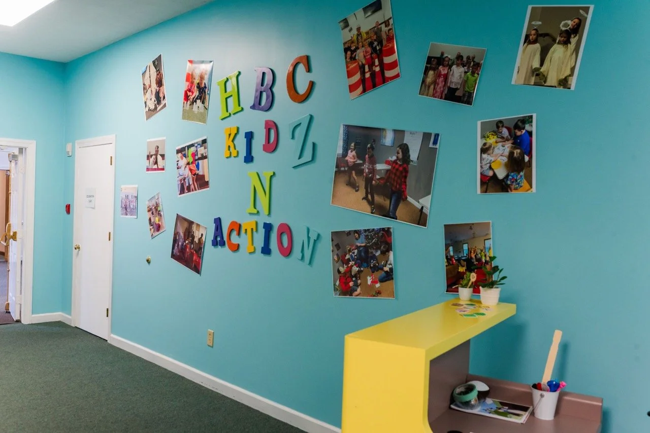 A woman reading a book to a group of children sitting on the floor outside, near a brick wall and framed animal posters.