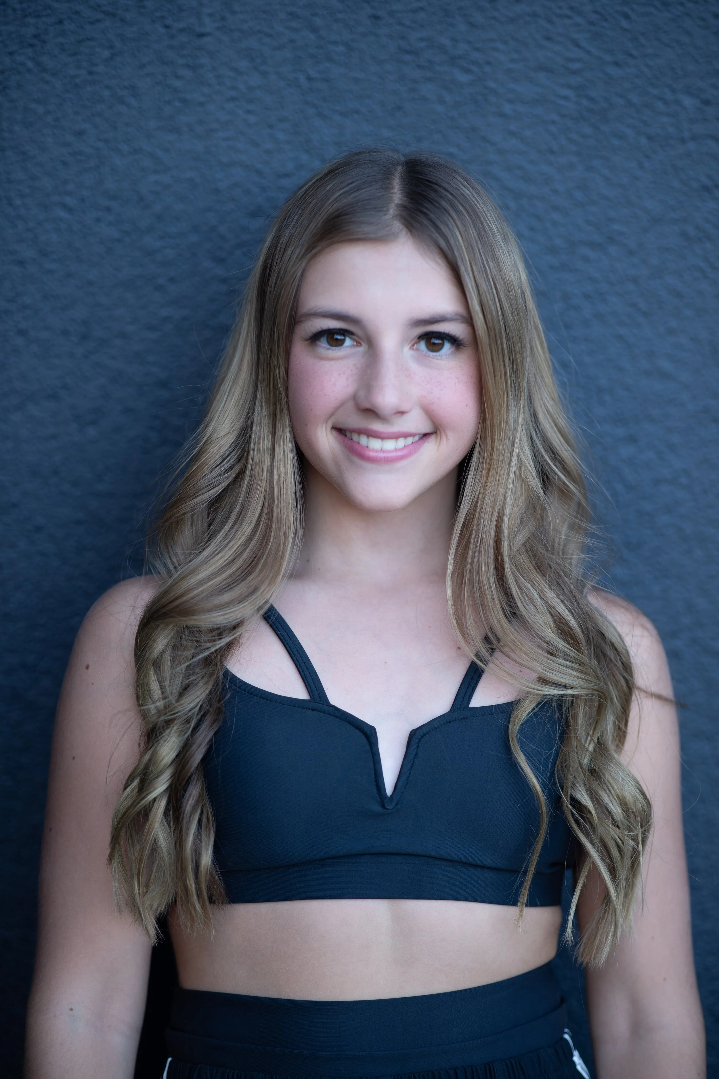 A young woman with long, wavy blonde hair wearing a black athletic top standing against a dark textured wall.