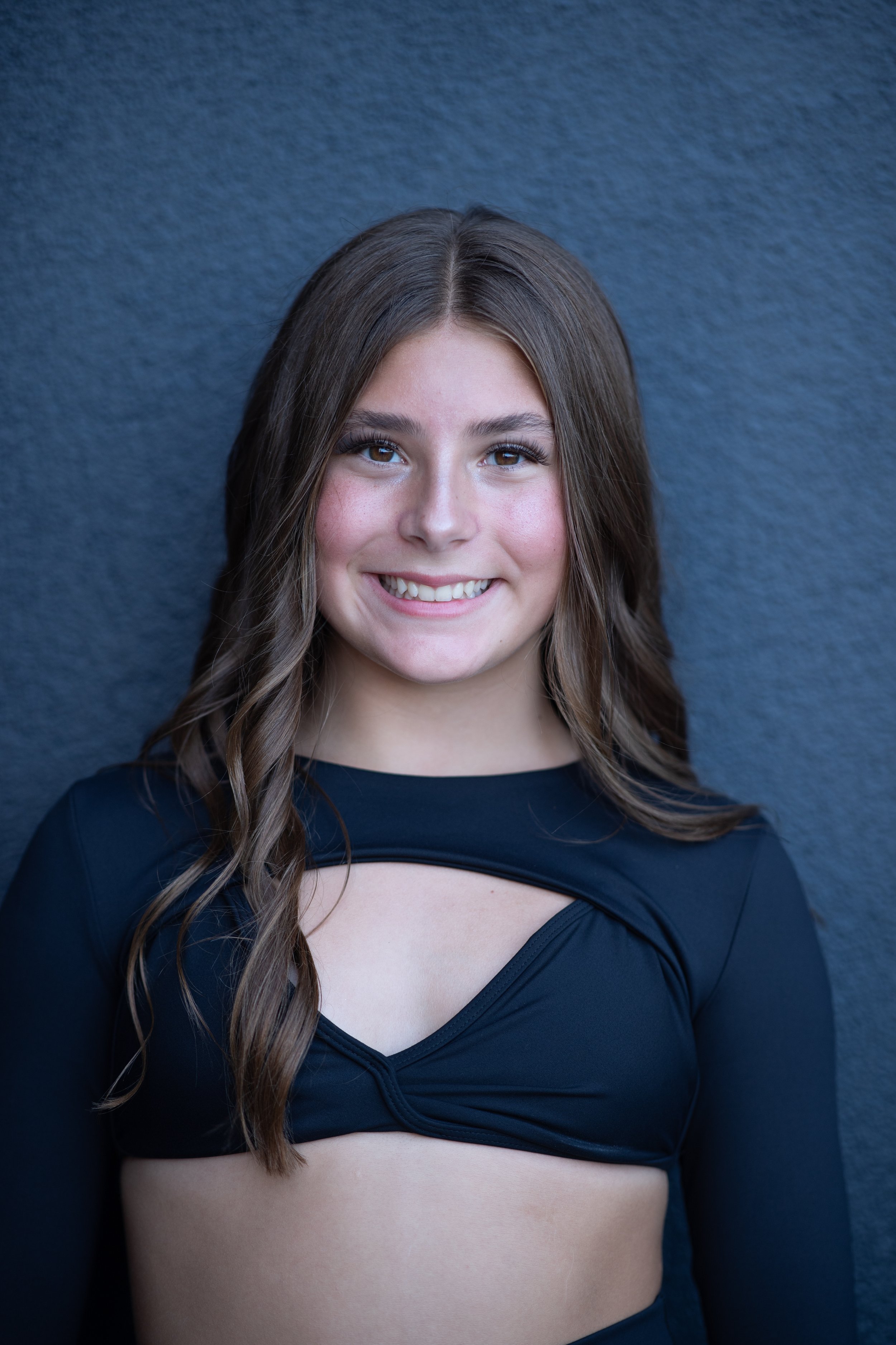 Portrait of a young woman with long wavy brown hair, smiling, wearing a black cut-out top, standing against a dark textured background.