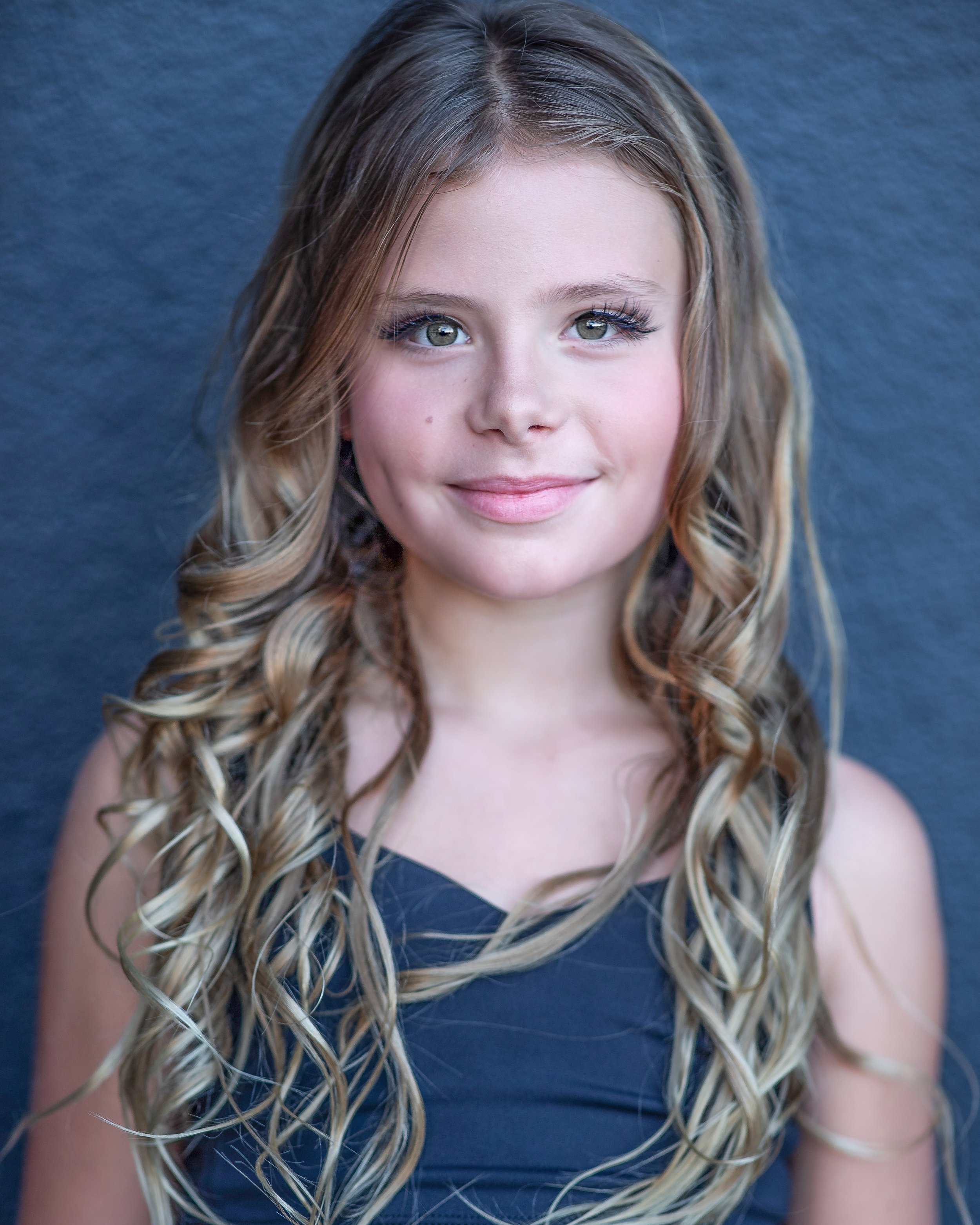 A young girl with long, wavy hair standing against a solid dark background, smiling softly at the camera.