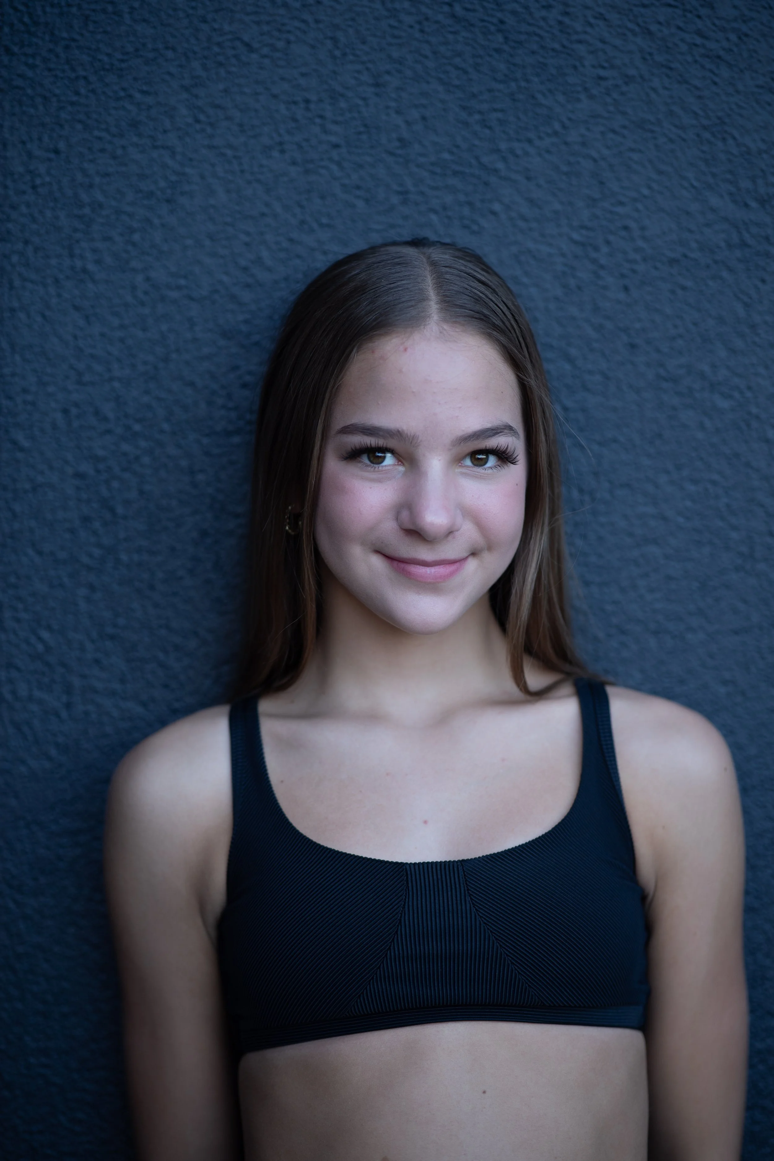 A young girl with long brown hair smiling, wearing a black athletic top, standing against a dark textured wall.