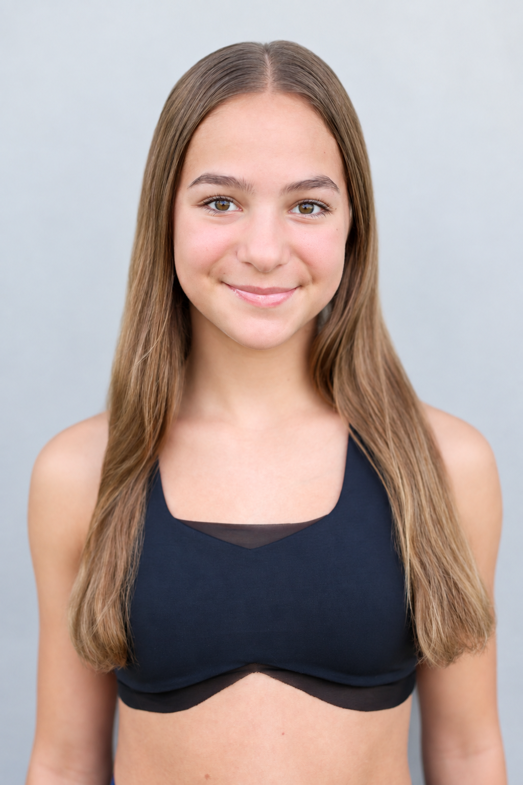 A young girl with long brown hair smiling, wearing a black athletic top, standing against a dark textured wall.