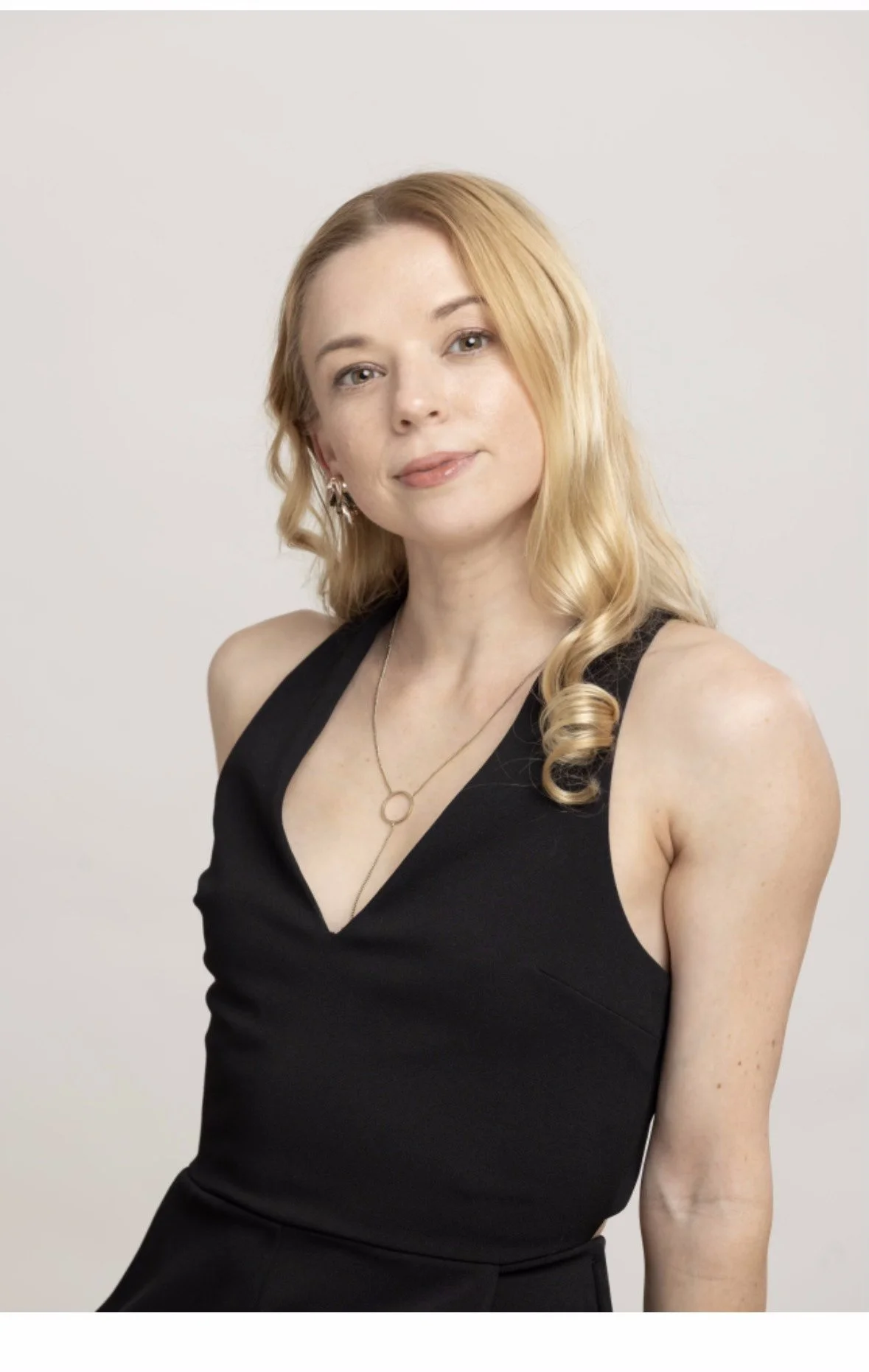 A woman with long blonde hair styled in loose curls wearing a sleeveless black dress and silver jewelry, posing against a plain light background.