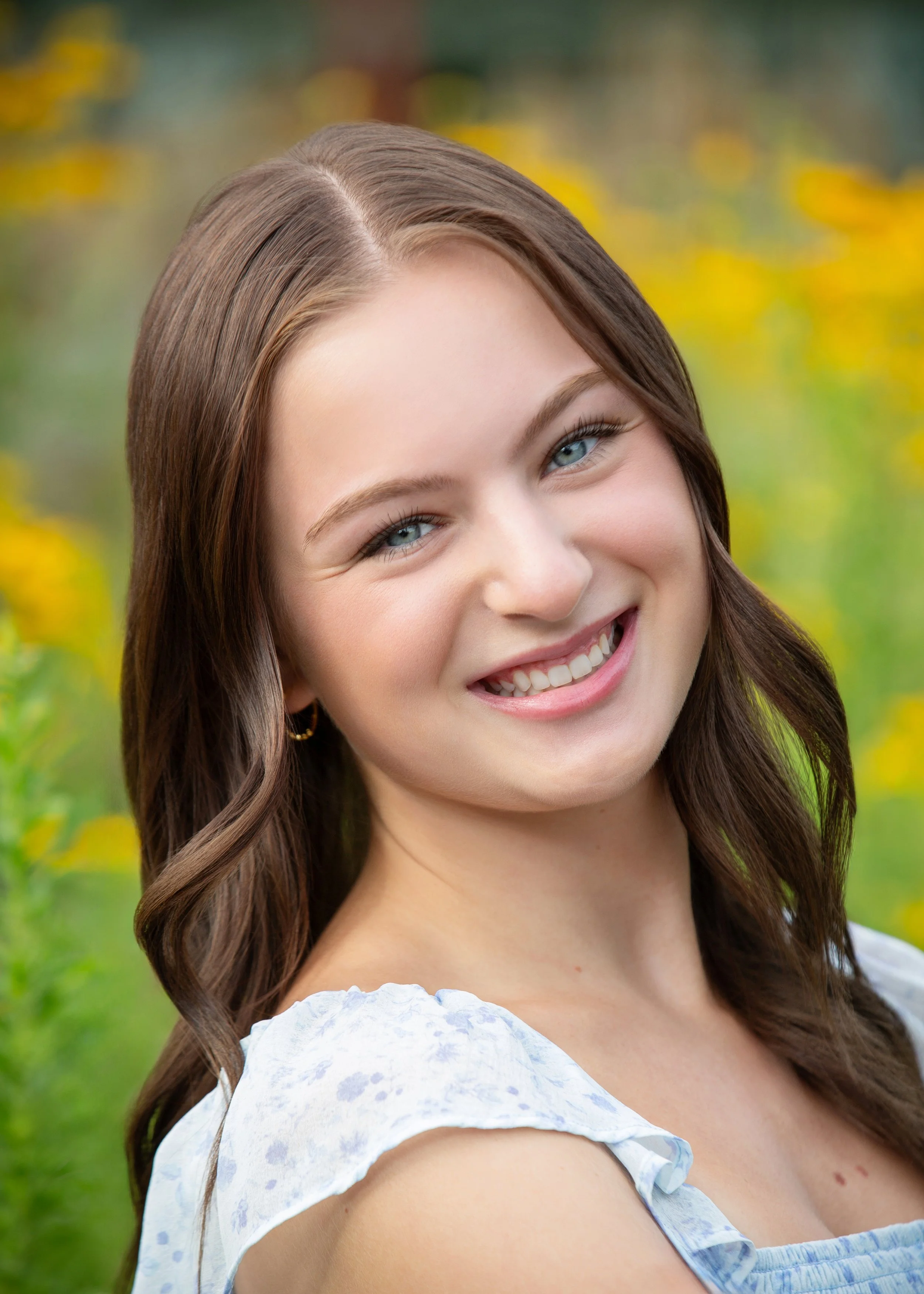 Close-up of a smiling young woman with long brown hair and blue eyes, wearing a light blue floral dress, outdoors with a blurred yellow and green background.