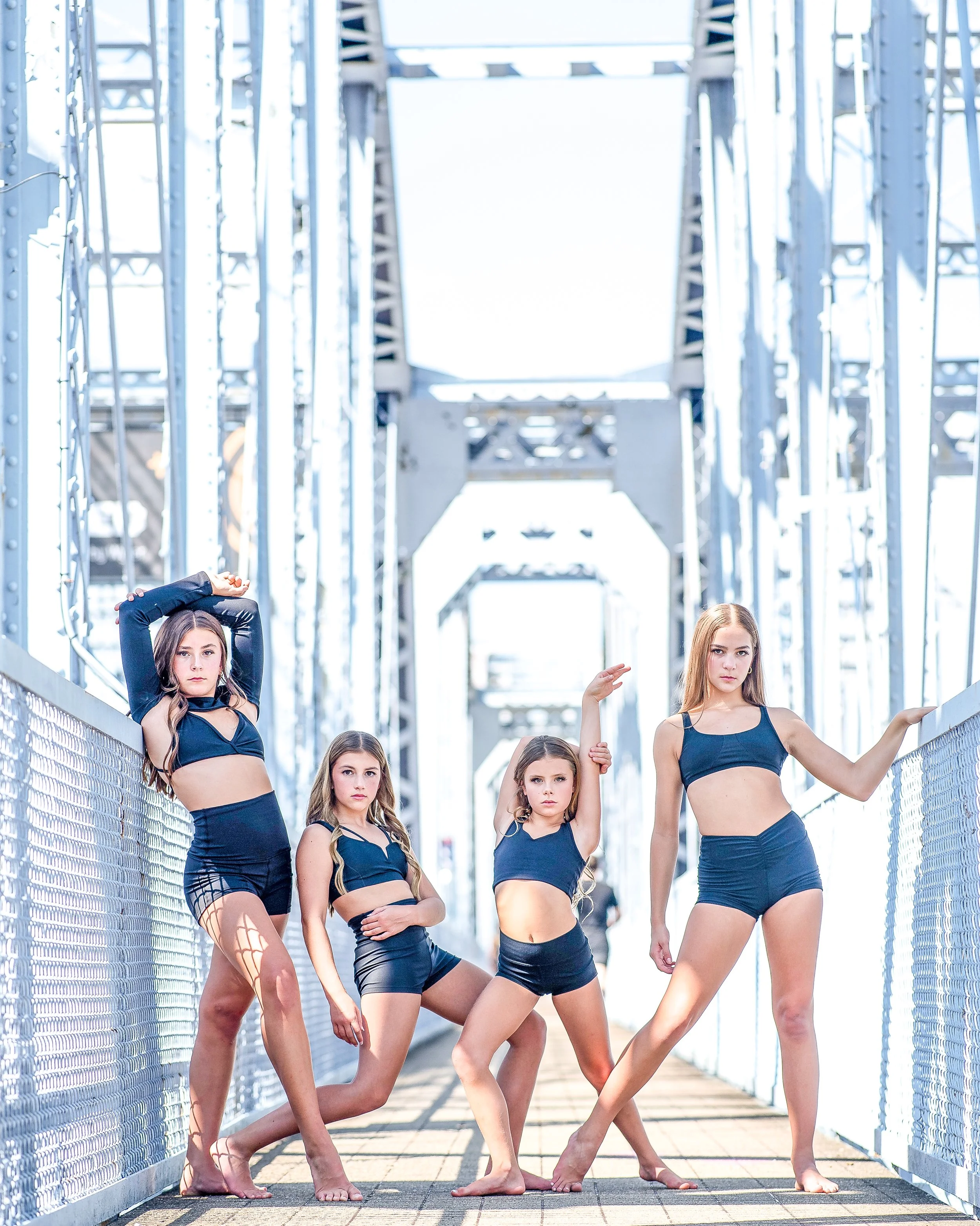 Four young women in black sports bras and shorts posing on a metal bridge with a bright sky background.