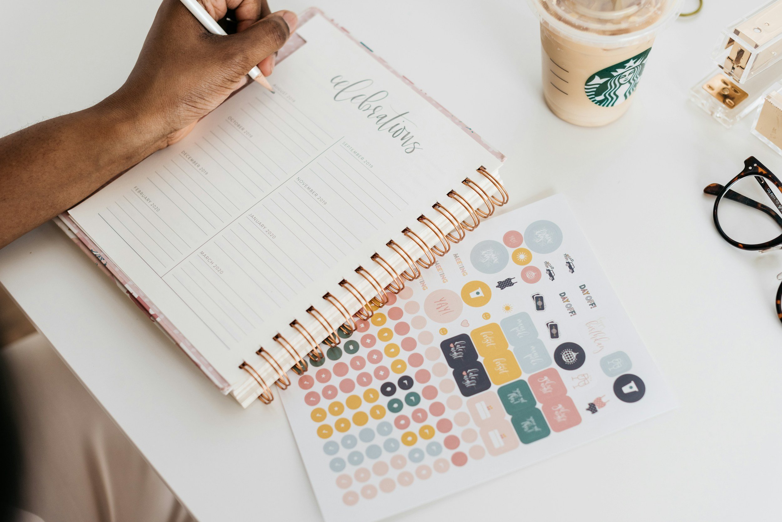 A person's hand is writing in a planner titled 'Celebrations' with sections for each month, on a white desk. There is a Starbucks iced coffee, a pair of glasses, and decorative stickers with various icons and words like 'yay' and 'happy' nearby.