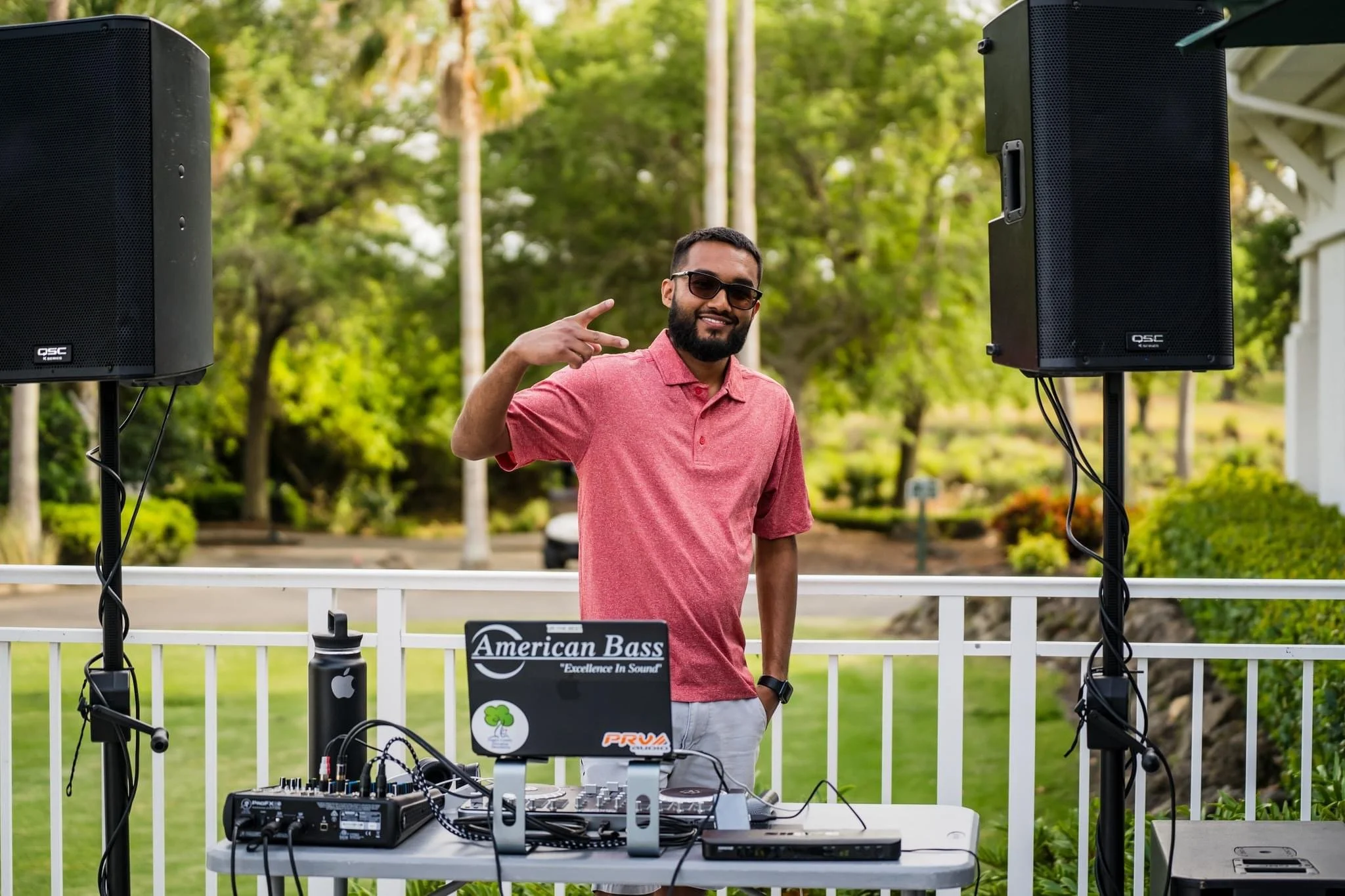 A man wearing sunglasses and a pink polo shirt standing outdoors behind DJ equipment, making a peace sign with his right hand, on a sunny day with trees and greenery in the background.