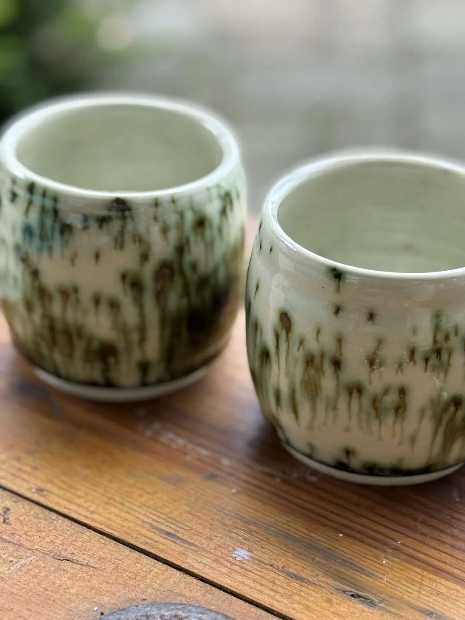 Two ceramic tea cups with green glaze and drip patterns, placed on a wooden surface outdoors.