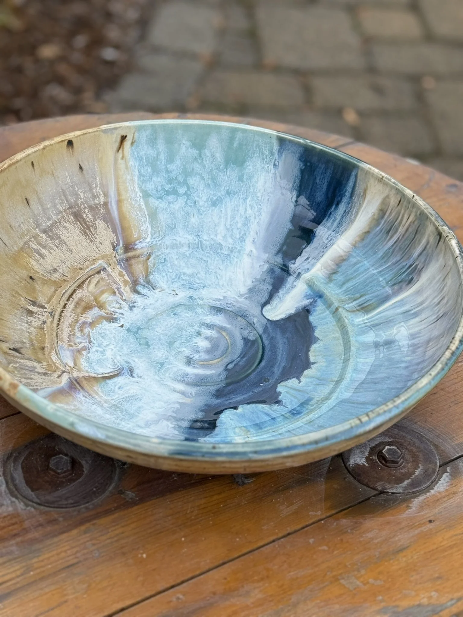 Colorful ceramic bowl with abstract blue, white, and beige glaze design, placed on a wooden surface outside.