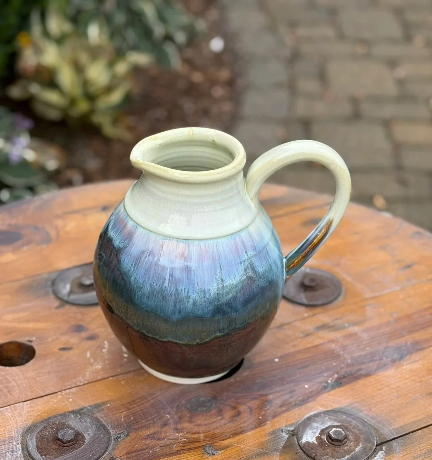 Colorful ceramic pitcher with a cream-colored neck and handle, placed on a wooden surface outdoors.