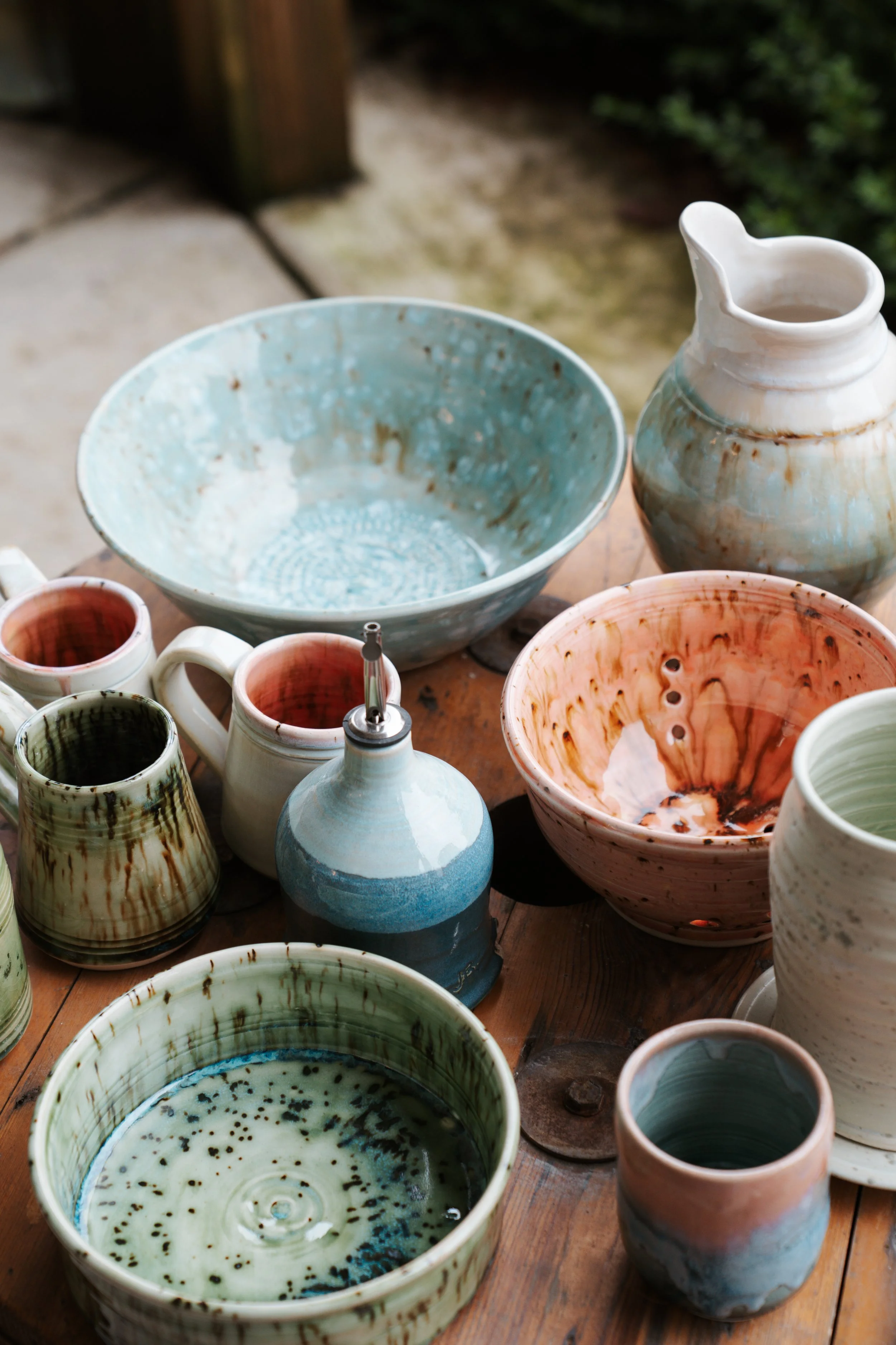 Various handmade ceramic bowls, mugs, and pitchers on a wooden table outdoors.