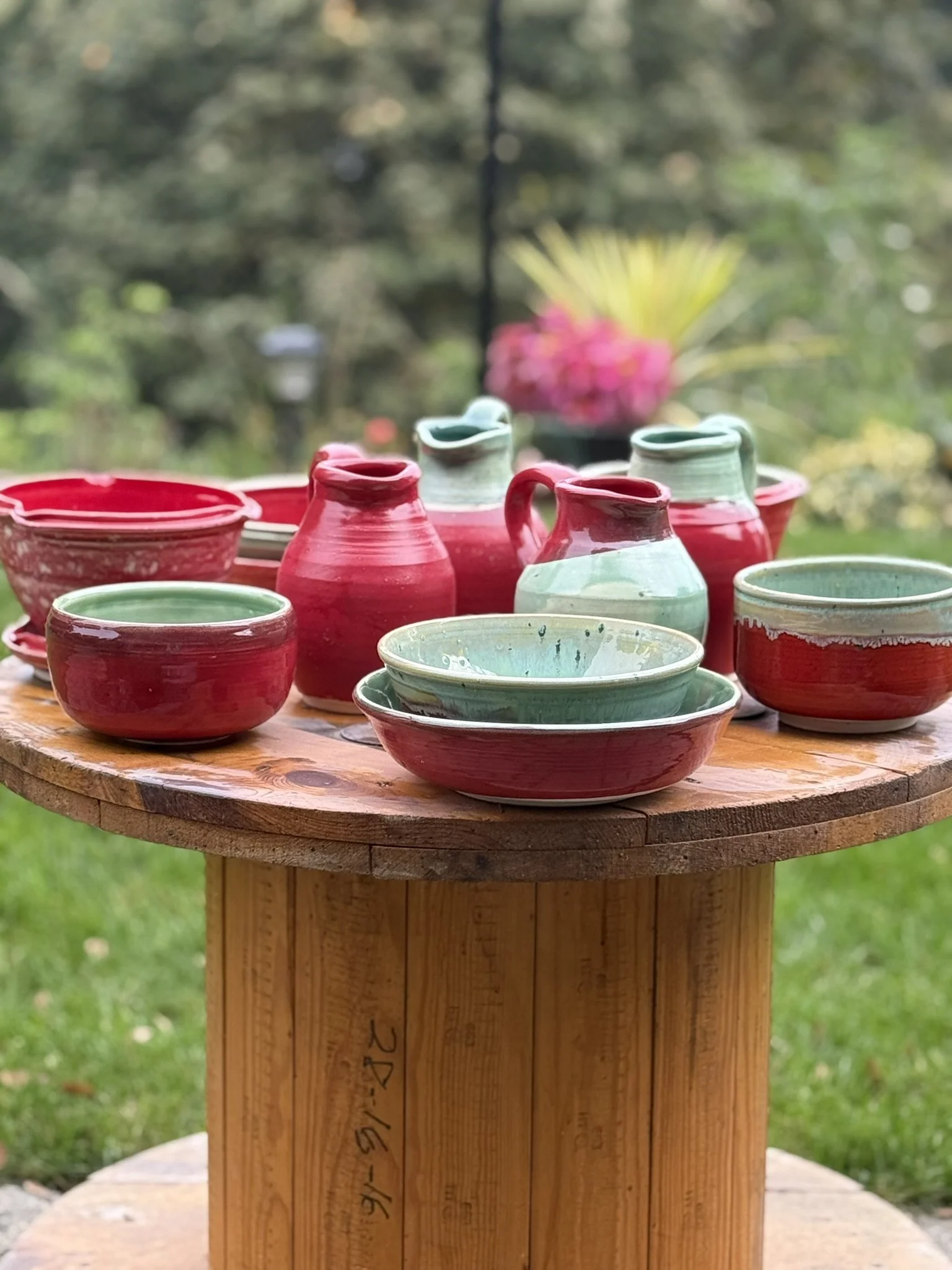 Colorful glazed pottery bowls, cups, and pitchers arranged on a round wooden table outdoors with a blurred garden background.