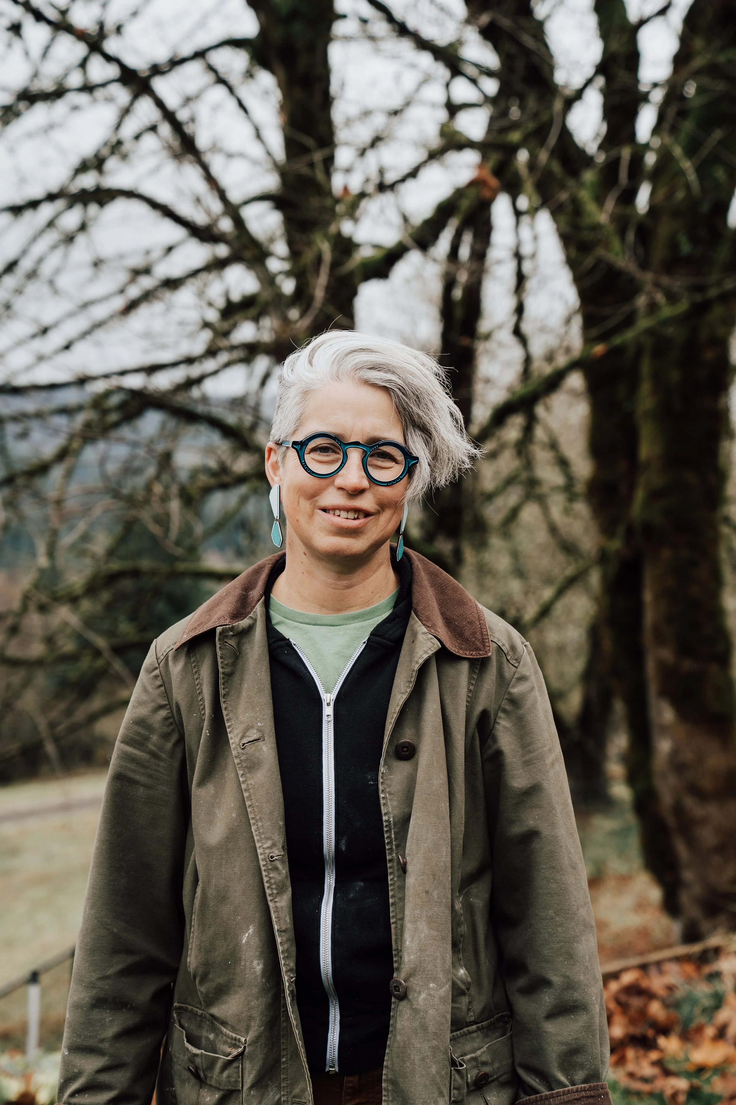 A woman with short gray hair, wearing glasses with round frames, earrings, and a green jacket over a black hoodie, standing outdoors in front of leafless trees.