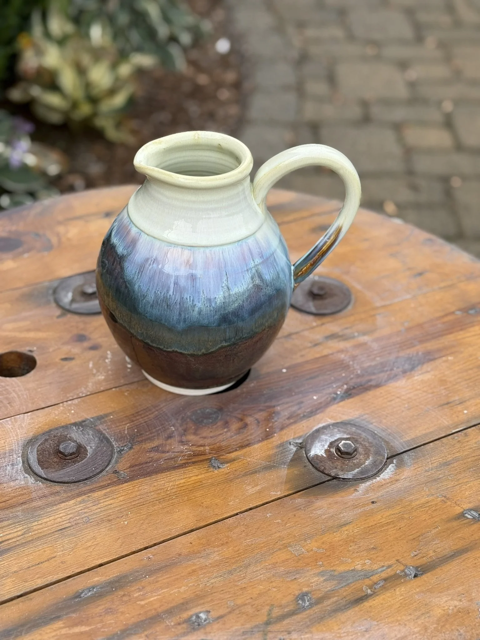 A ceramic pitcher with a landscape glaze, sitting on a wooden outdoor table with bolts, with garden plants and brick pathway in the background.