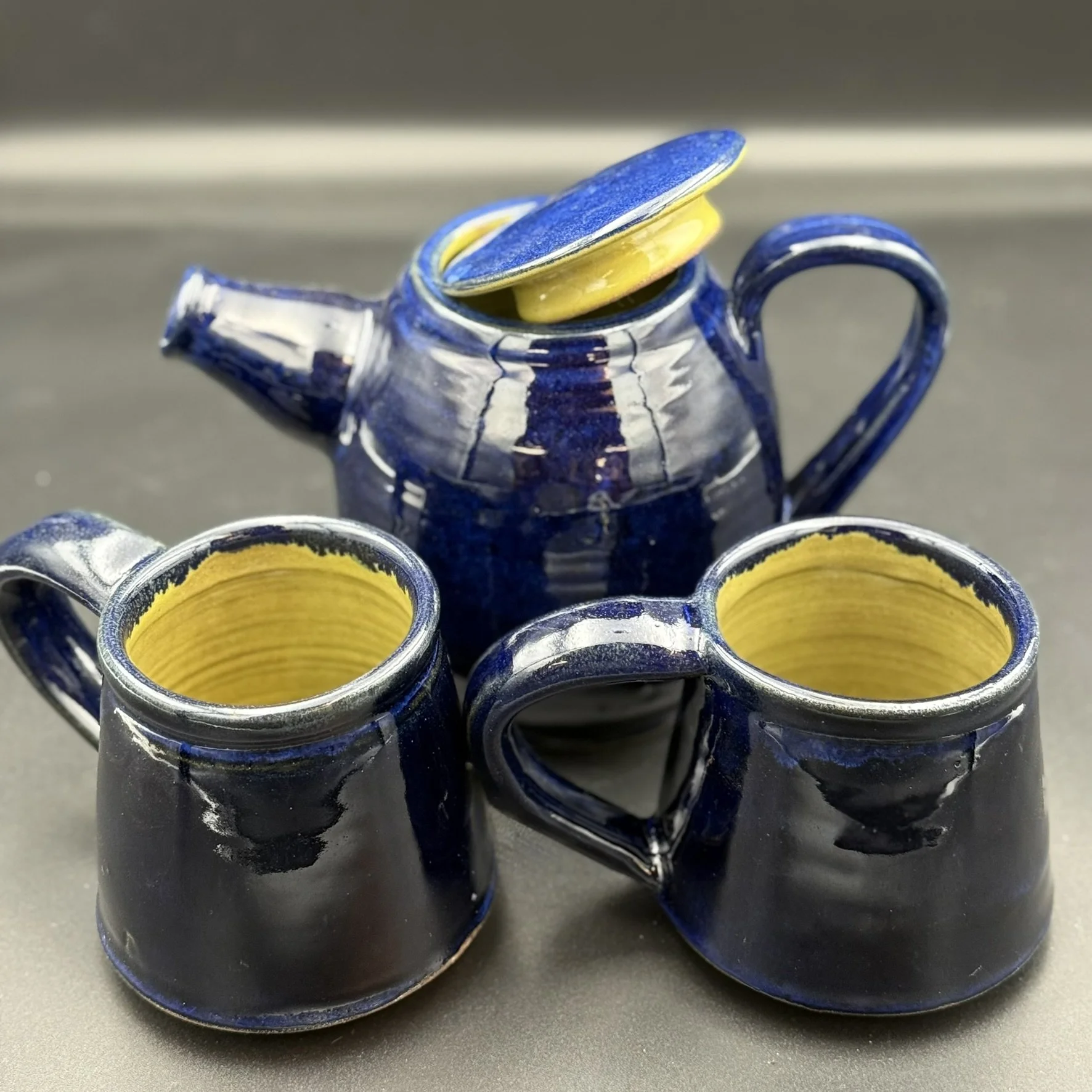 A small blue ceramic teapot with a yellow interior, with yellow and blue ceramic cups stacked inside and two blue cups in front with yellow interiors.