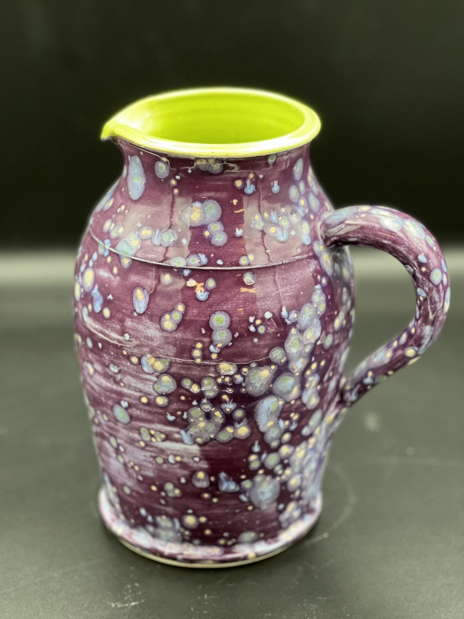 A ceramic pitcher with a purple and white bubble pattern, a green interior, and a curved handle, placed on a dark surface.
