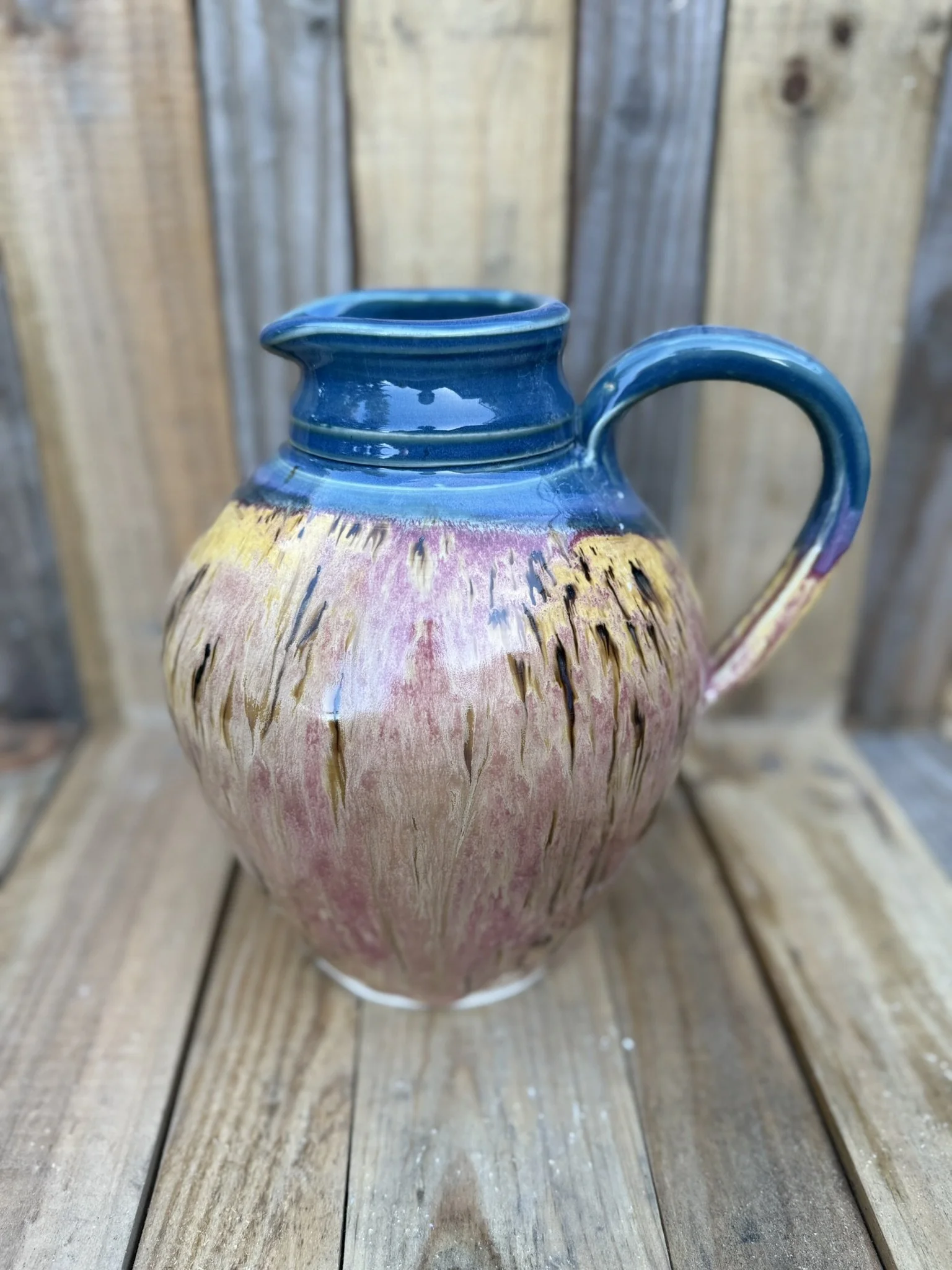 Colorful ceramic jug with a rounded body, blue rim and handle, and a glaze with pink, yellow, and purple streaks, placed on a wooden surface with a wooden fence background.