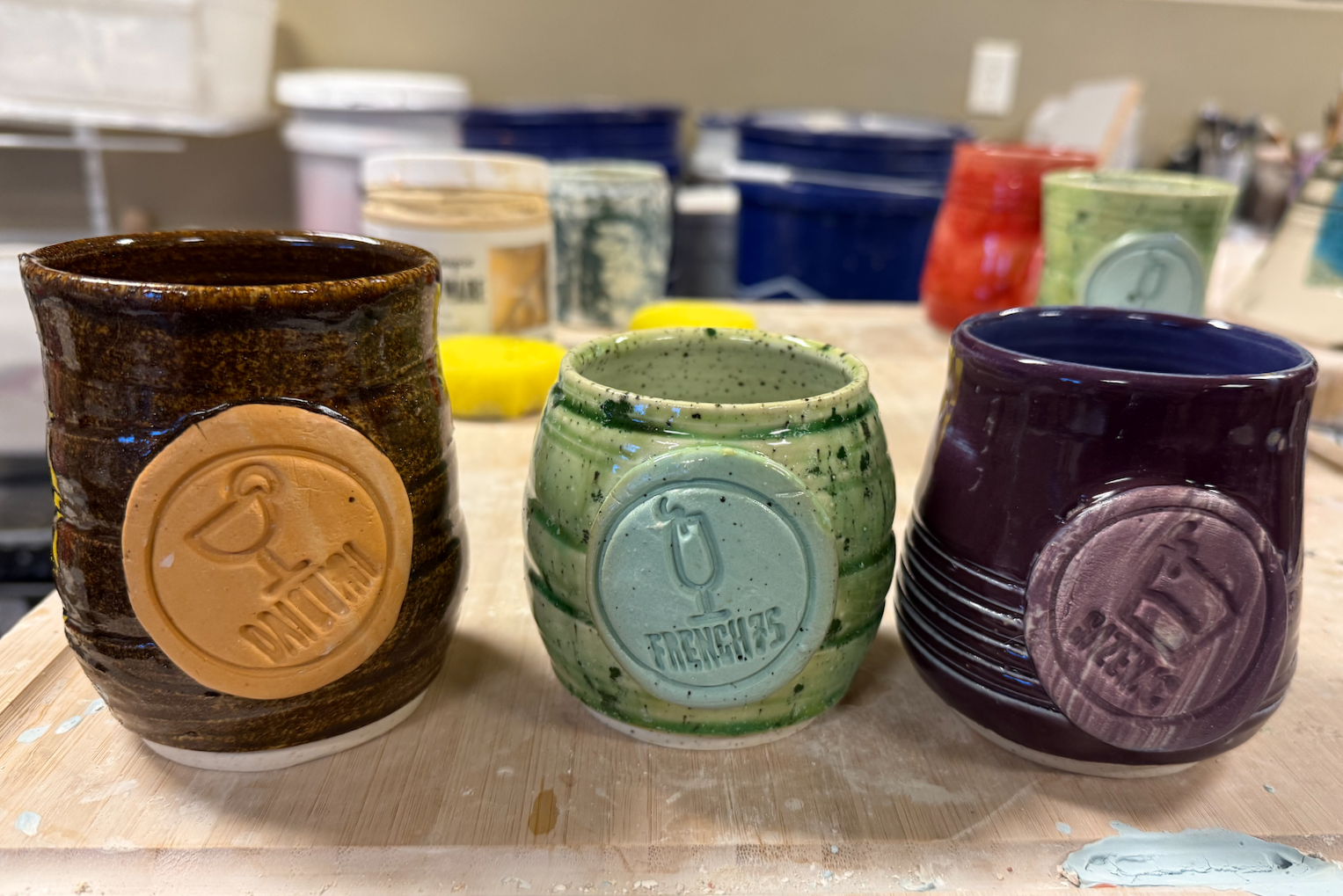 Three glazed ceramic mugs with embossed beer logos on a work table in a pottery studio.