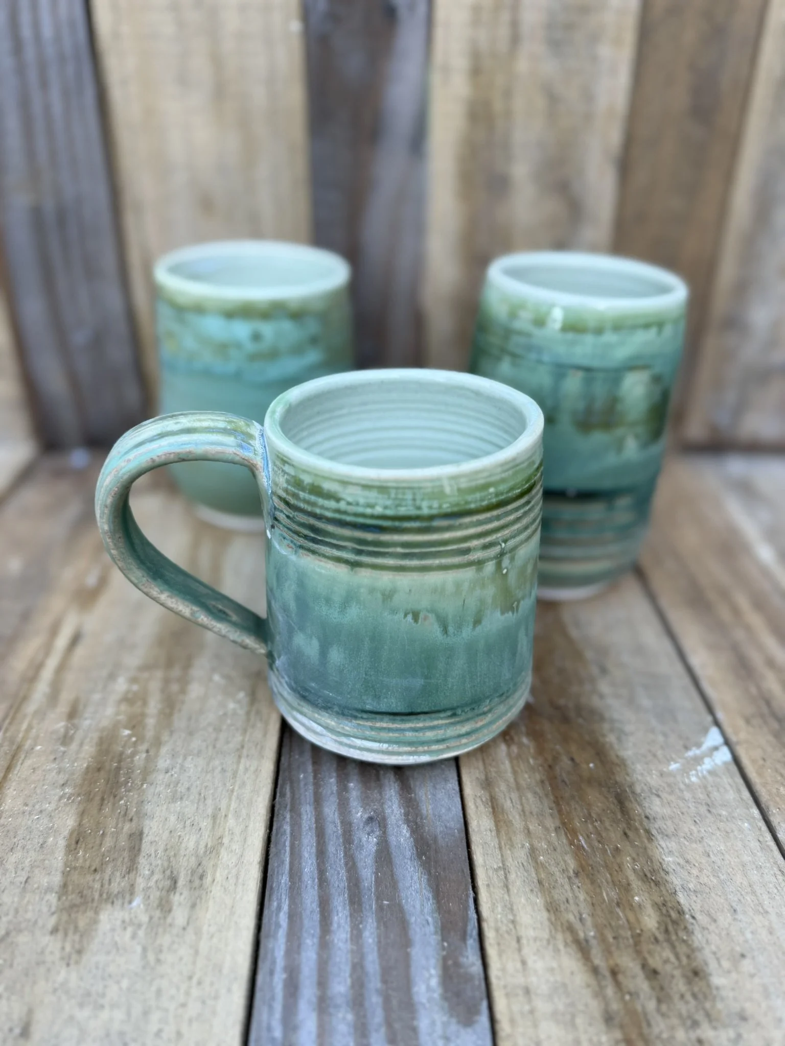 Three ceramic mugs with a green and blue glaze, placed on a wooden surface with a wooden background.