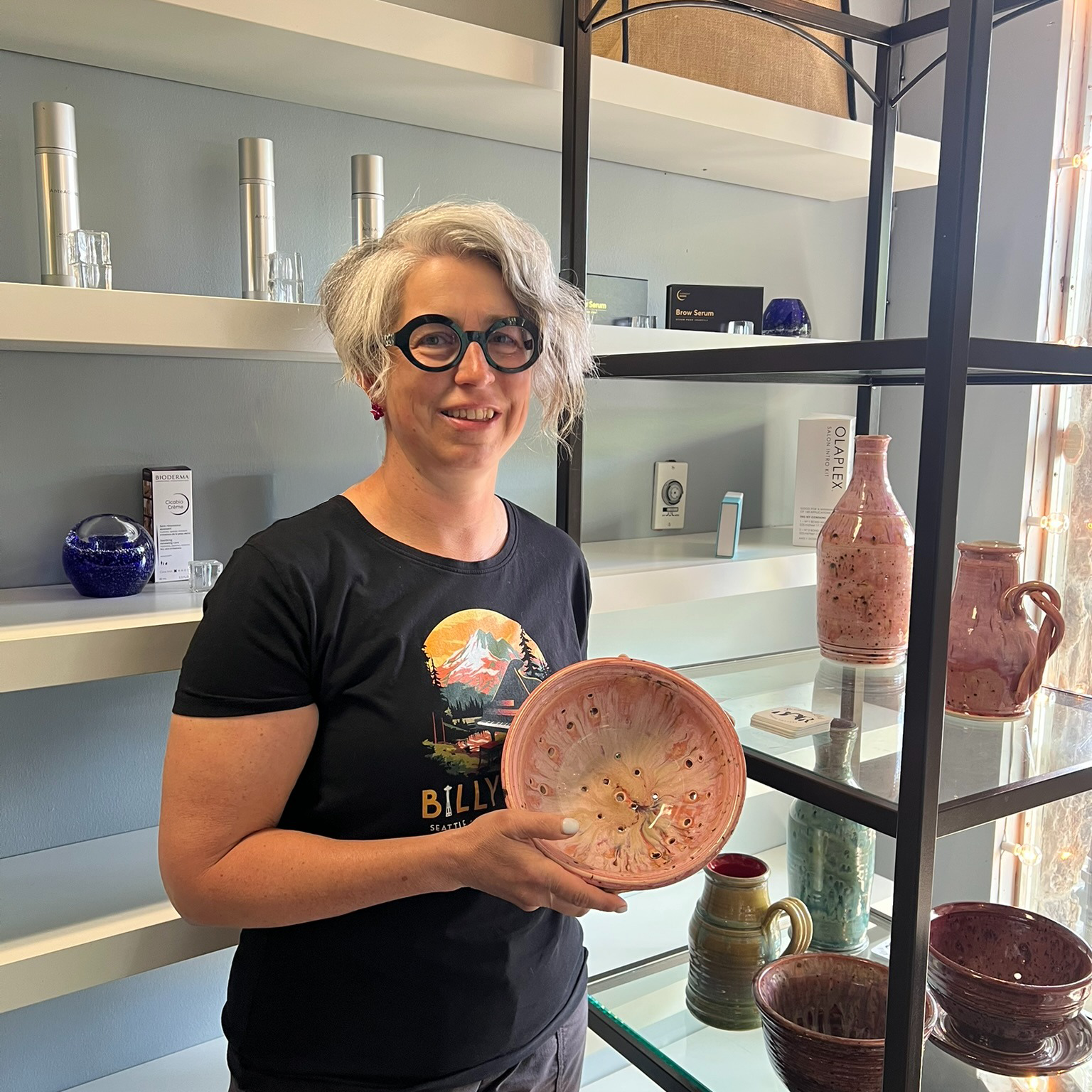 A woman with gray curly hair, glasses, and earrings is holding a pink, glazed ceramic bowl and standing in front of shelves displaying various pottery pieces, including vases and bowls, in a pottery shop.