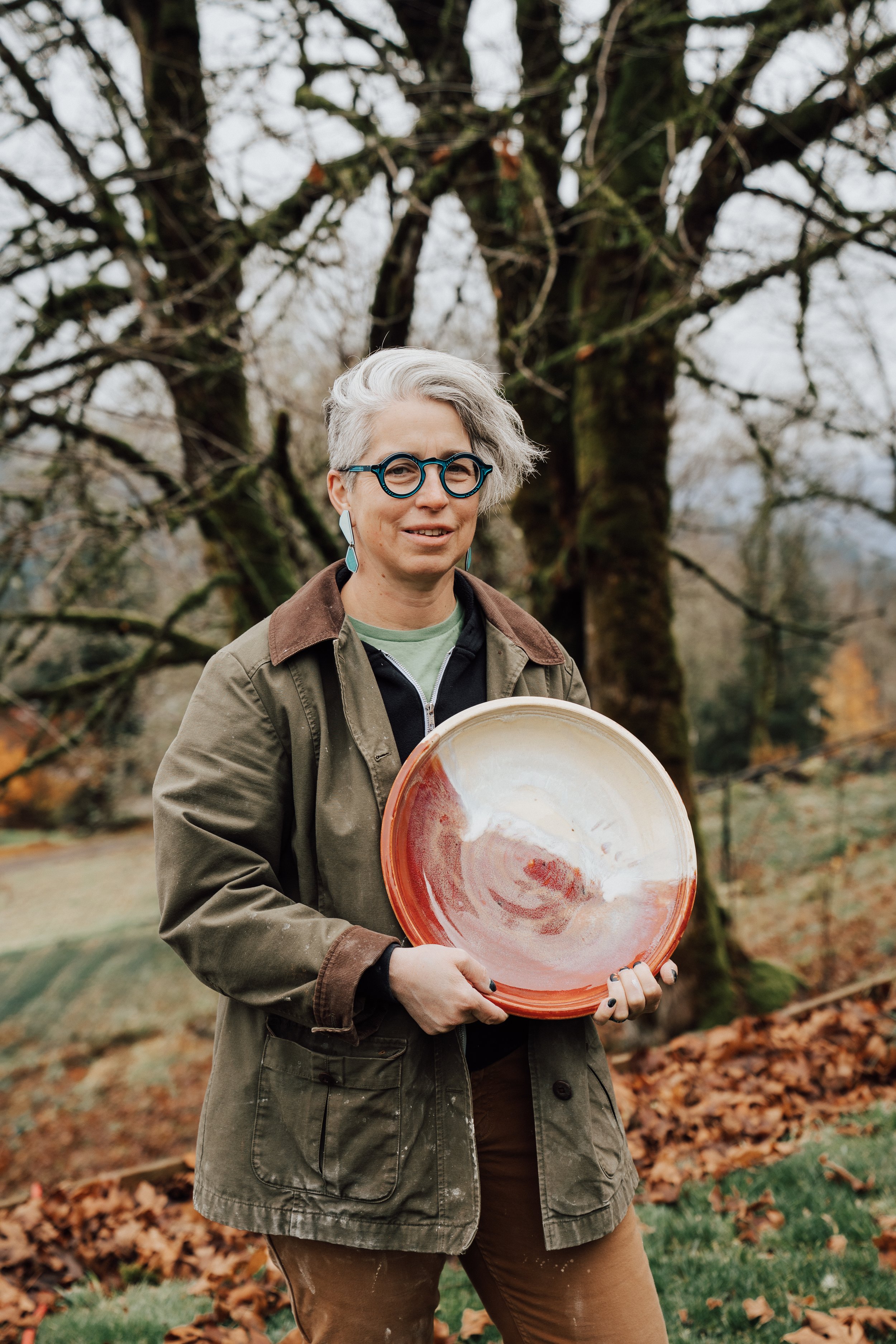 A woman with short gray hair and round glasses holding a large ceramic plate outdoors. She is wearing a green jacket with brown accents, a black shirt, and brown pants, standing in front of a tree with bare branches and fallen leaves on the ground.