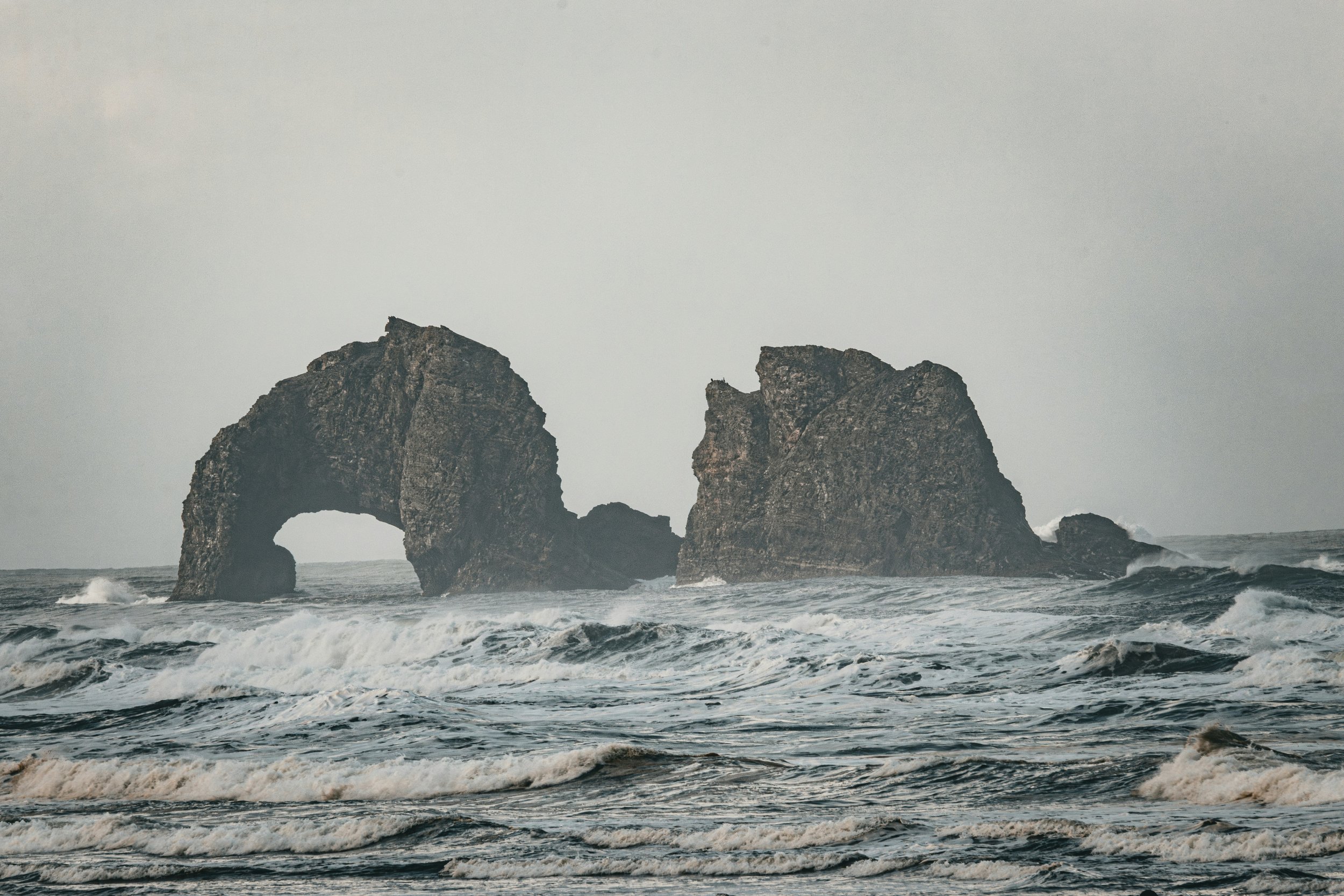 Two large sea stacks, one with a natural arch, rising from the ocean with waves crashing around them under an overcast sky.