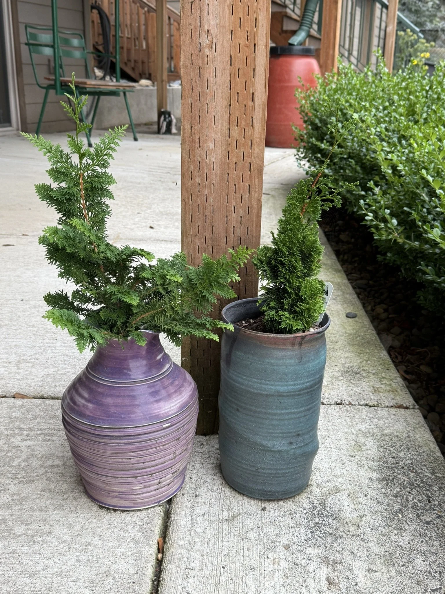 Two potted plants with small green trees are placed on a concrete porch next to a wooden post. One pot is purple with horizontal ridges, and the other is blue-green with a smooth surface. In the background, there is outdoor furniture and a red water 