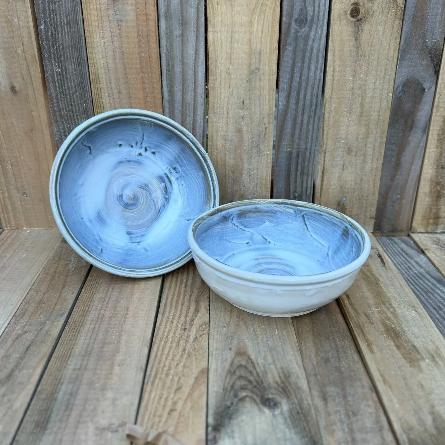 Two ceramic bowls with blue and white swirled glaze, placed on a wooden surface against a wooden fence background.