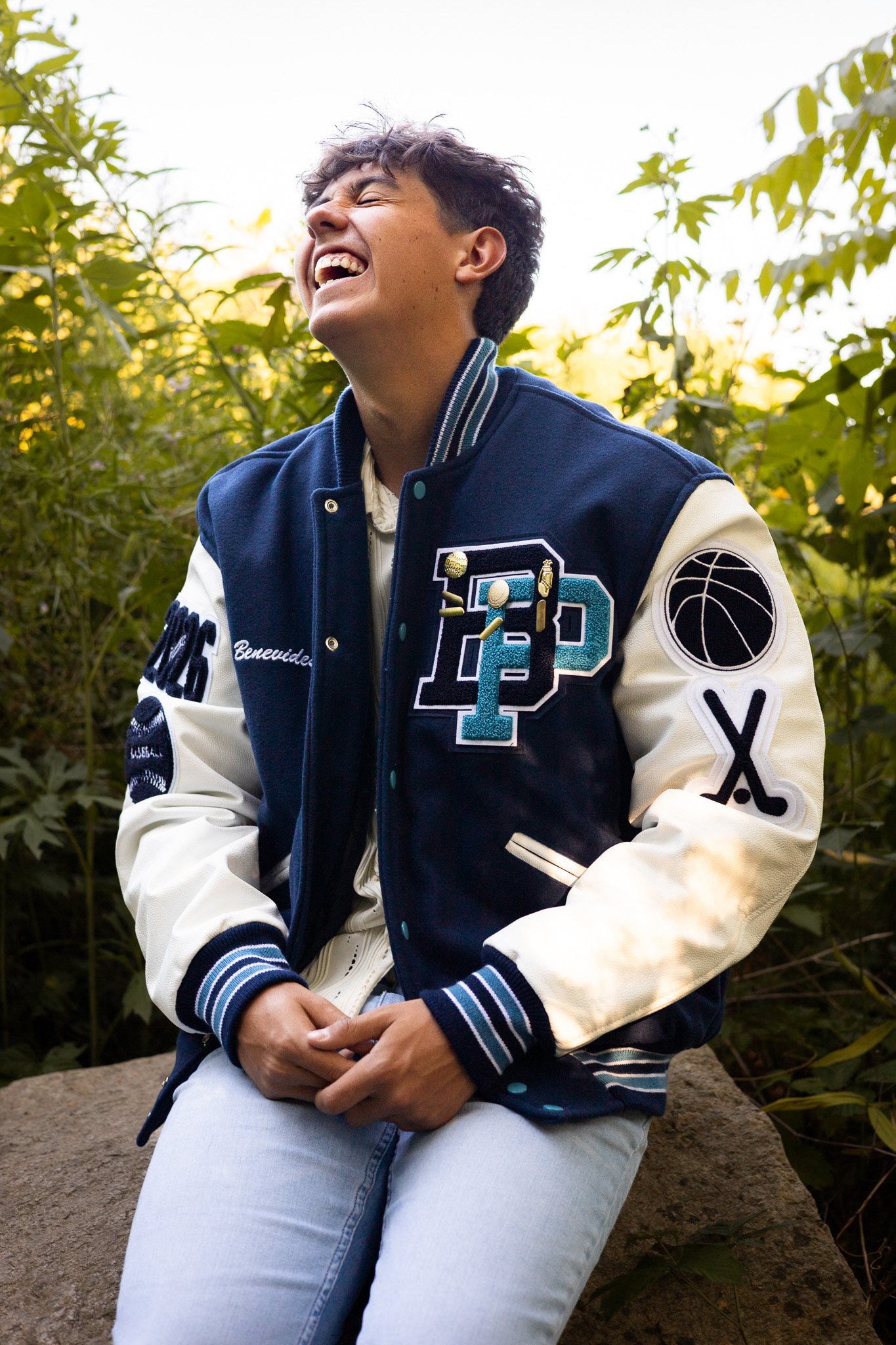 A young man sitting on a large rock, laughing and smiling, wearing a navy and cream varsity jacket with patches, light blue jeans, and a white shirt, surrounded by green plants and trees outdoors during daytime.