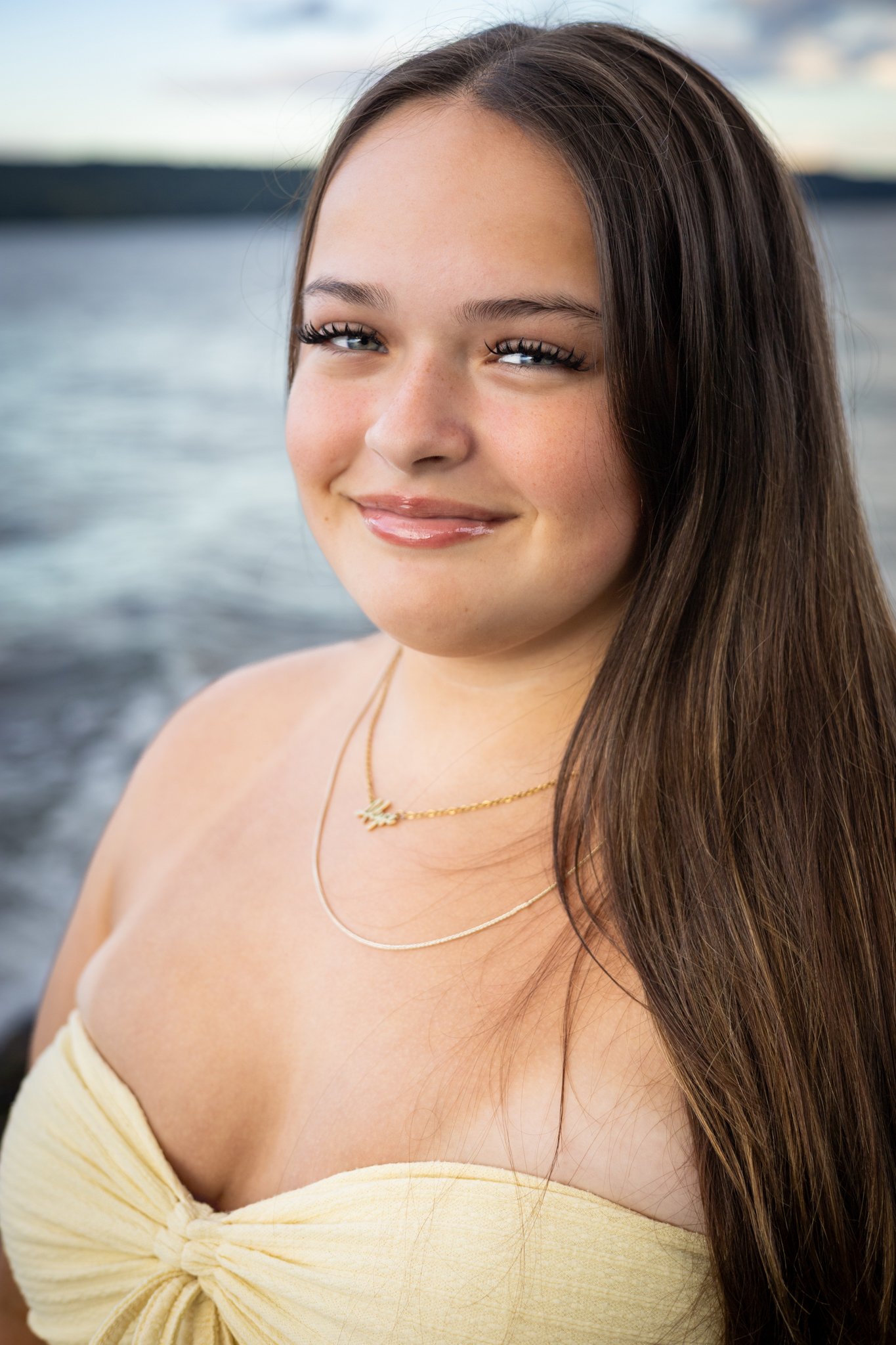 Close-up of a young woman with long brown hair, blue eyes, and a gentle smile, wearing a yellow off-shoulder top and gold jewelry, standing by a body of water during sunset.