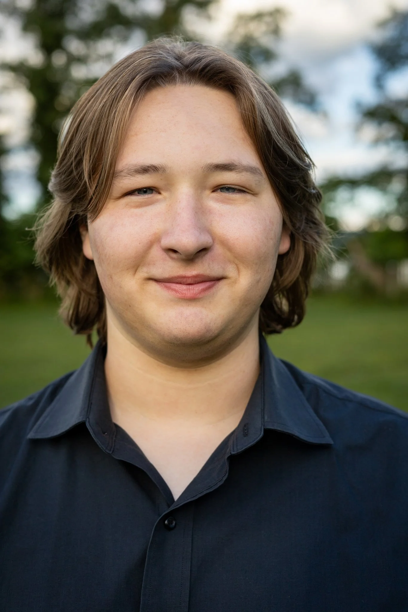 Close-up of a young person with light skin, brown hair, and blue eyes, smiling slightly, wearing a black button-up shirt, outdoors with blurred green trees and a partly cloudy sky in the background.