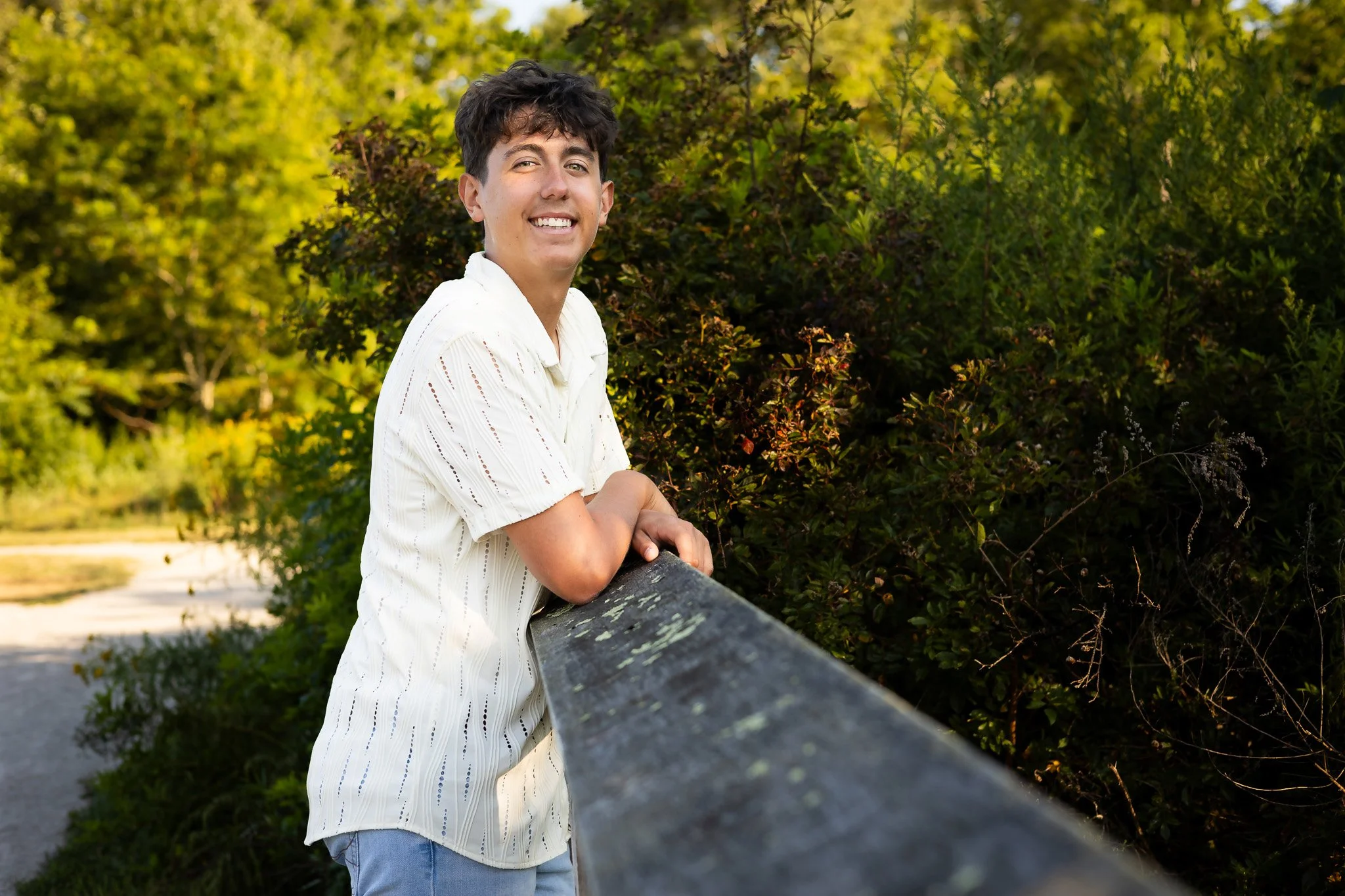 A young person with short dark hair wearing a white shirt and light blue jeans leaning on a wooden railing with lush green trees and bushes in the background.