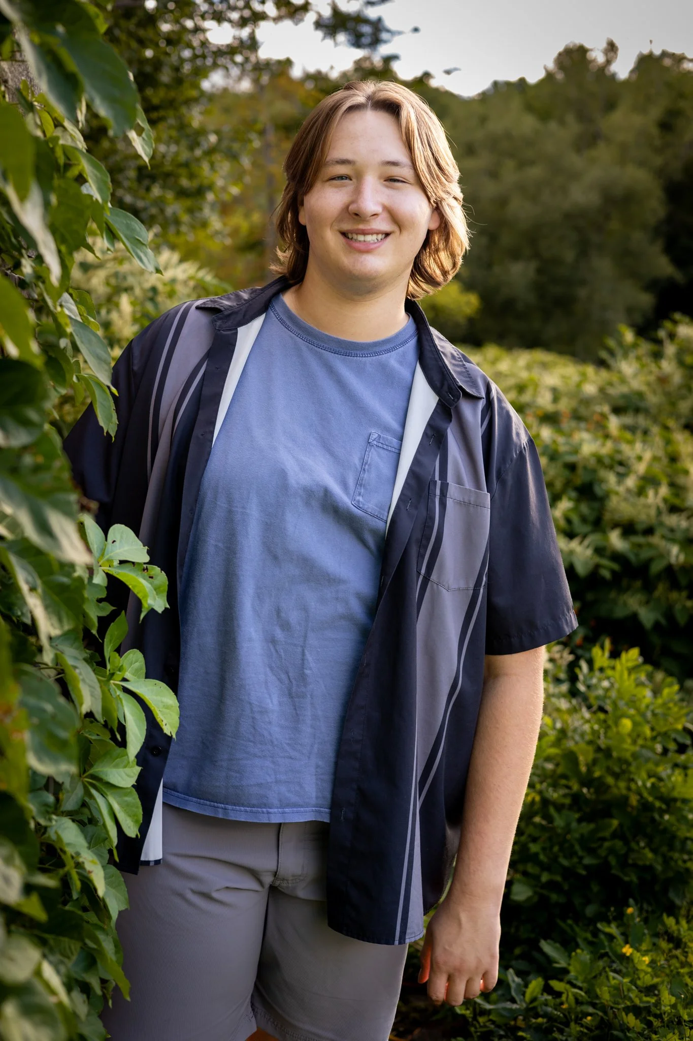 A young man with shoulder-length brown hair, smiling, standing outdoors in front of green trees and bushes, wearing a blue t-shirt and a dark jacket.